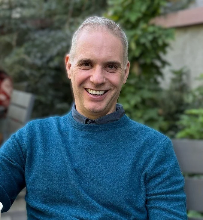 A smiling man with gray hair wearing a blue sweater outdoors with green foliage in the background.