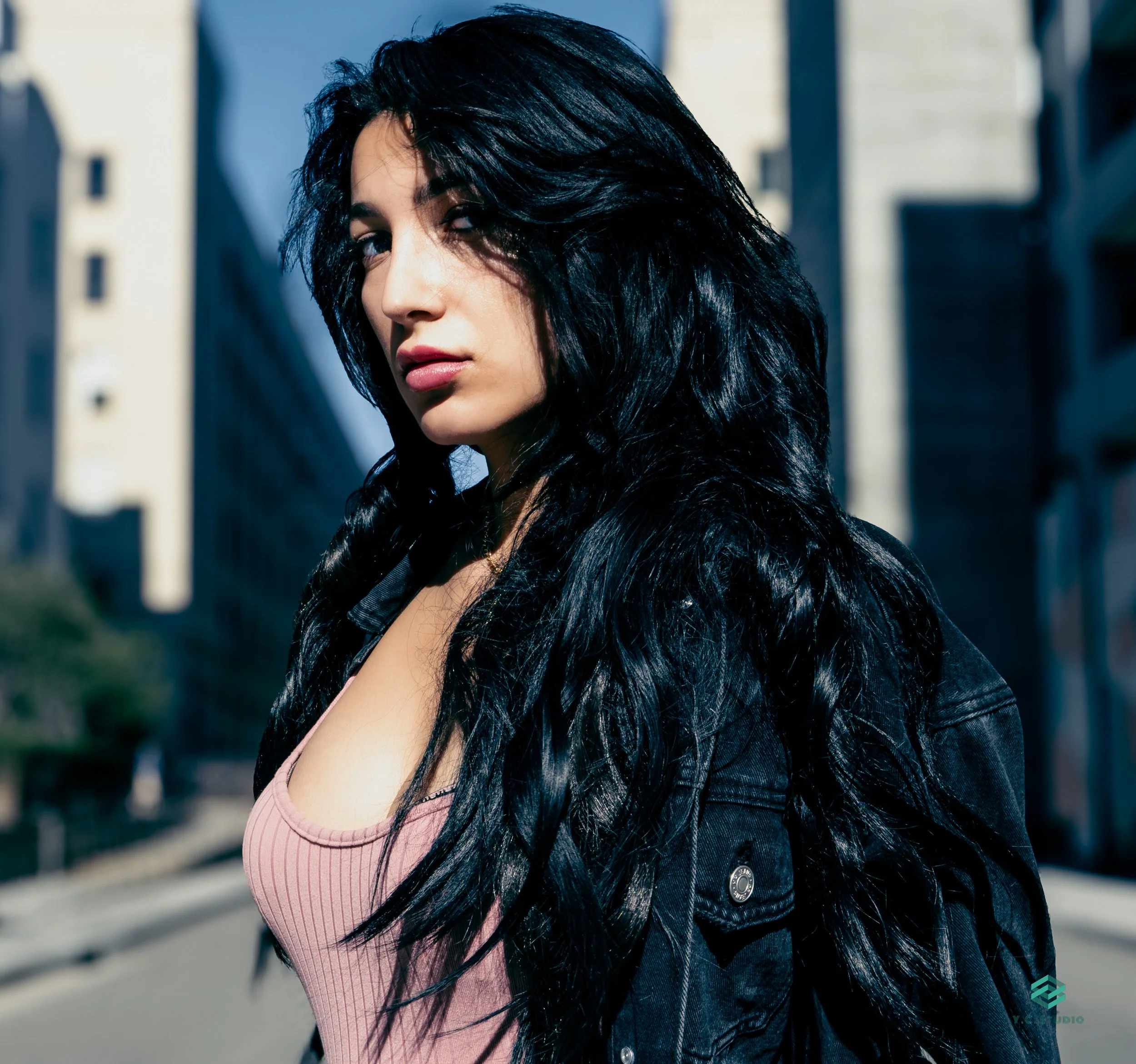 A young woman with long black curly hair, wearing a pink top and black jacket, standing outdoors in an urban area with buildings in the background.