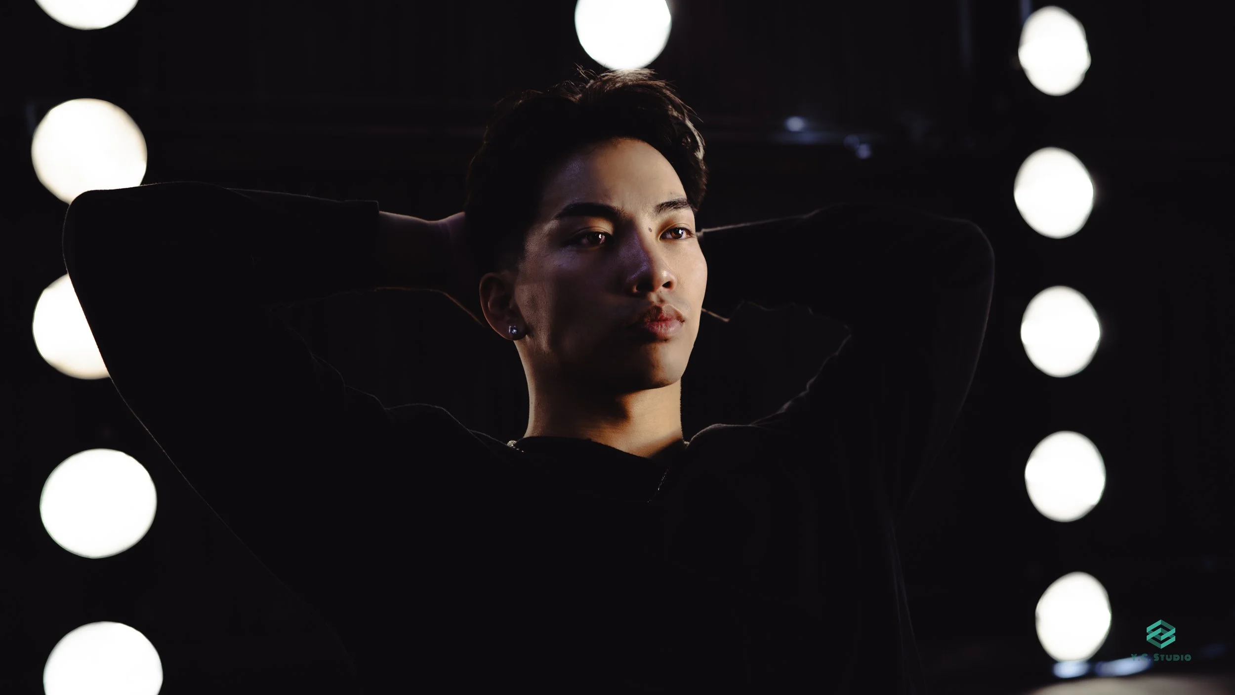 A man with dark hair posing with arms behind head in front of illuminated round lights in a dark setting.