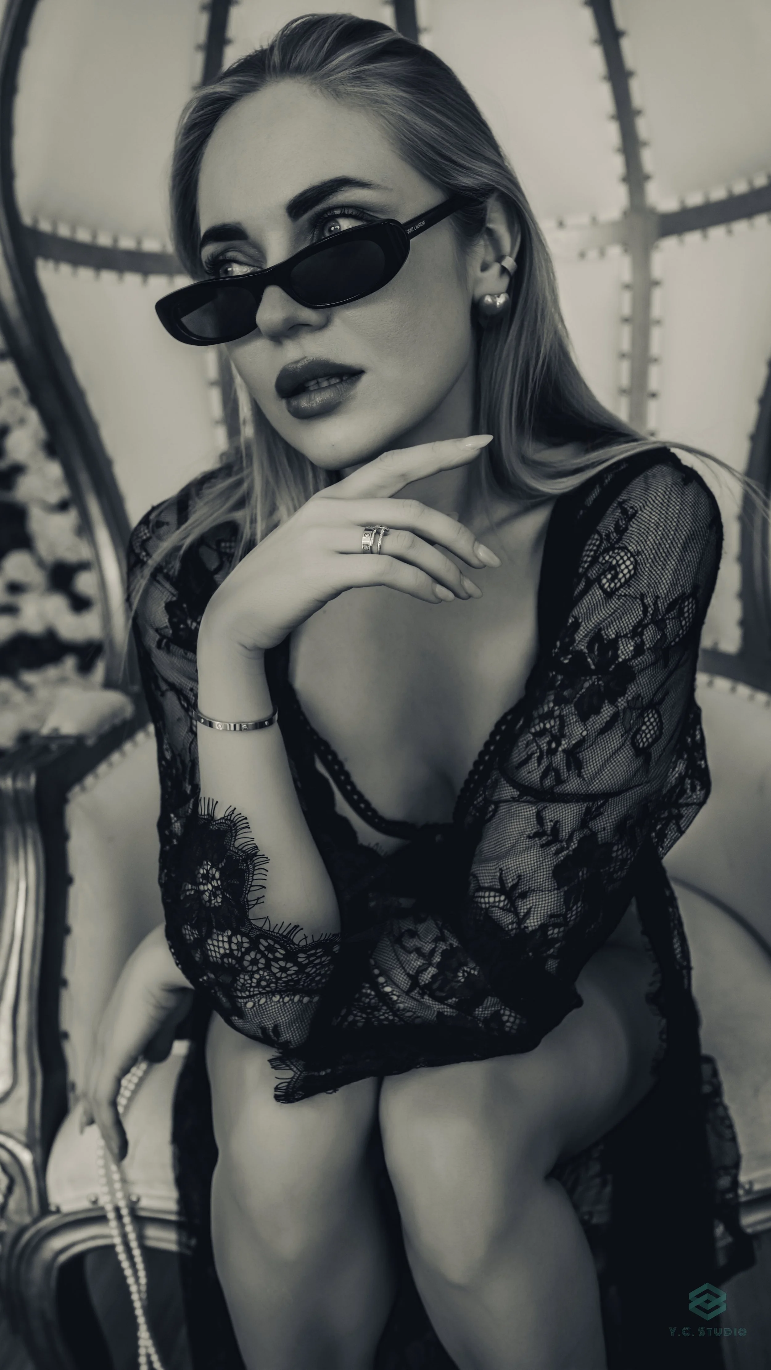 A woman with long hair, wearing dark sunglasses, a black lace top, and earrings, sitting on a vintage-style upholstered chair, with her hand near her face, in a black and white photograph.