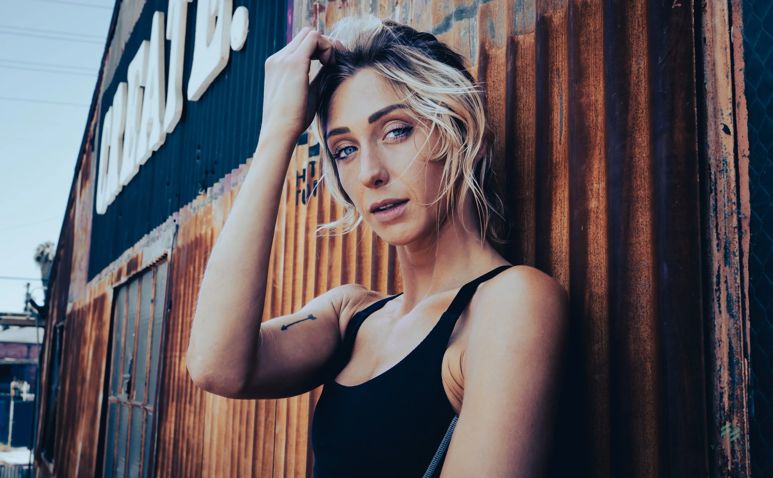 A young woman with blonde hair and blue eyes standing against a wooden wall. She is wearing a black sleeveless top and has a small tattoo on her left arm. She is touching her hair with her right hand and looking at the camera.