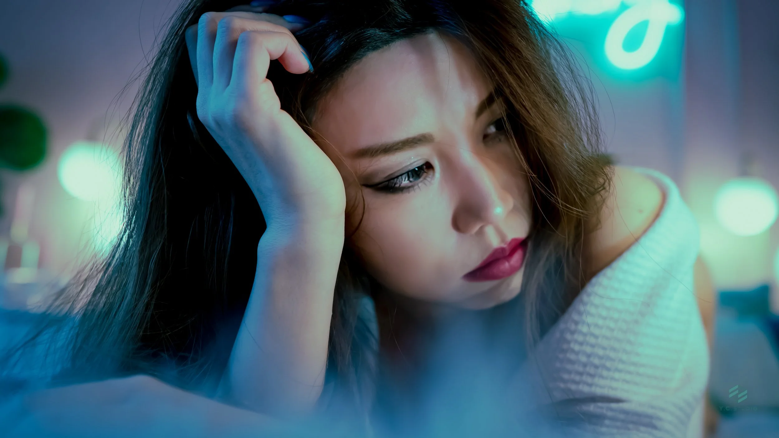 A woman with dark wavy hair and makeup, resting her head on one hand, appears thoughtful and introspective, in a setting with colorful neon lights.