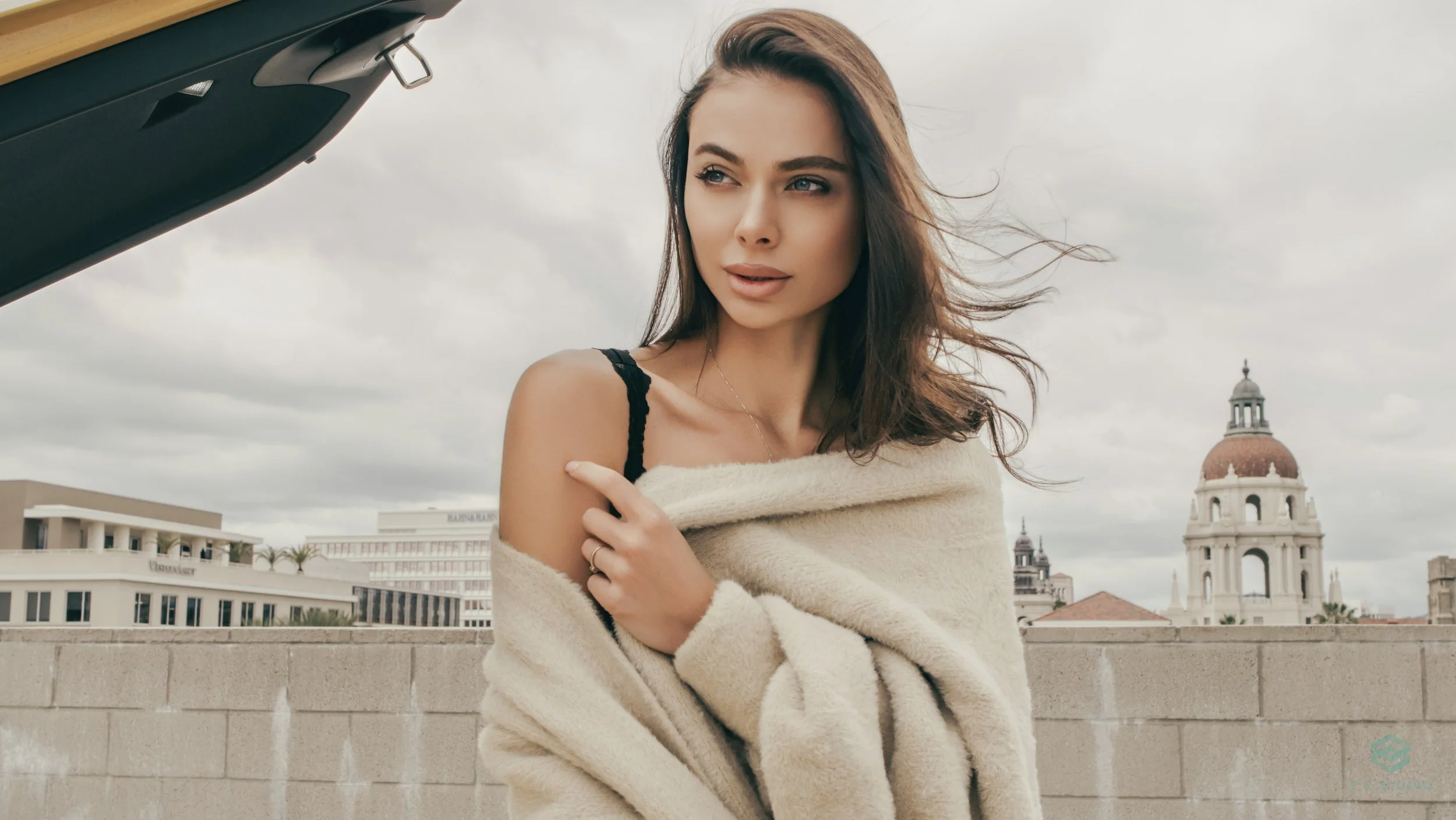 A young woman with long brown hair standing on a rooftop with city buildings and a domed tower in the background. She is wearing a black top, partially covered by a beige coat, and looks to the side with a contemplative expression.