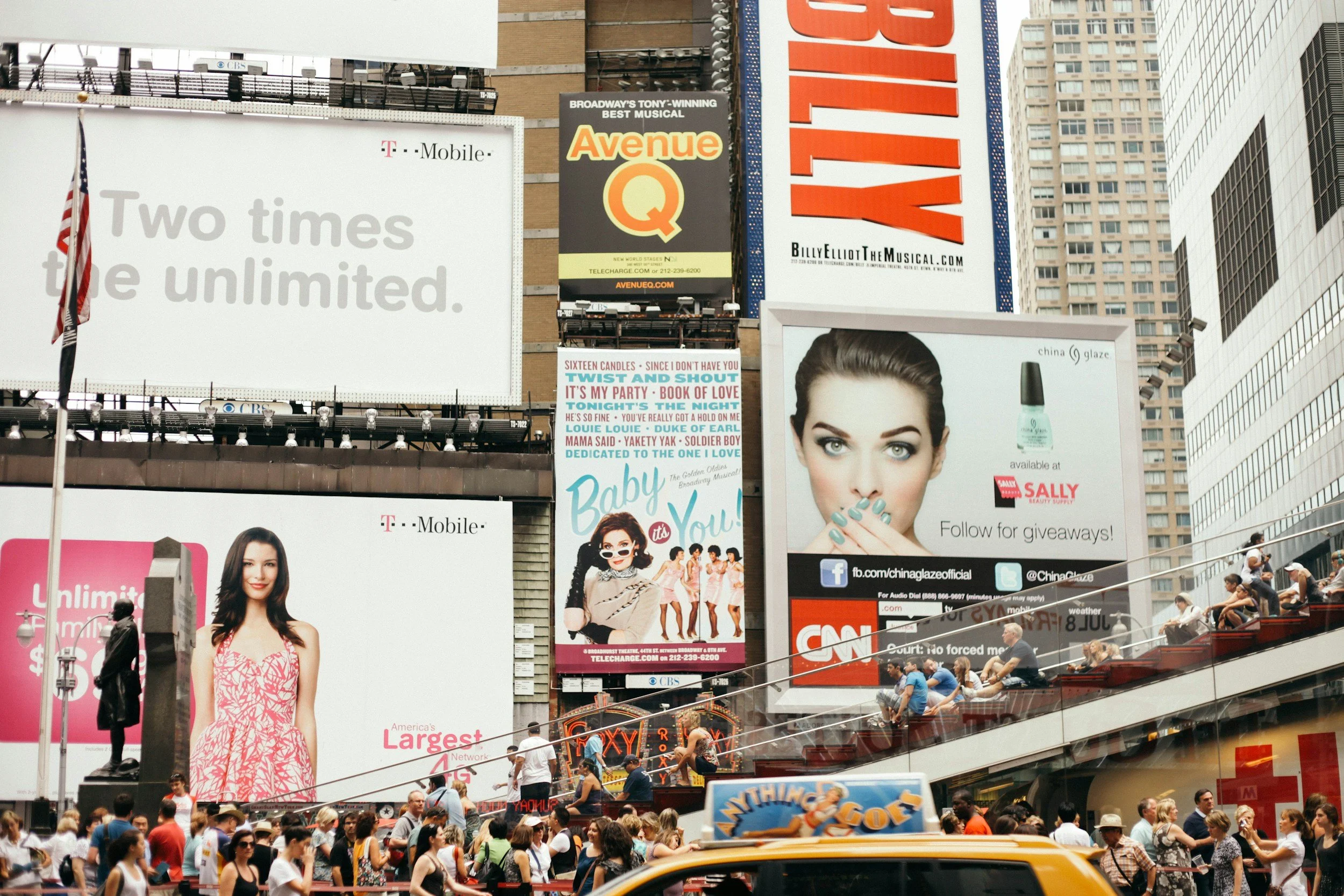 Crowd walking in Times Square, New York City, with large billboards and advertisements, including for T-Mobile, Avenue Q musical, and a beauty product, with a staircase and buildings in the background.
