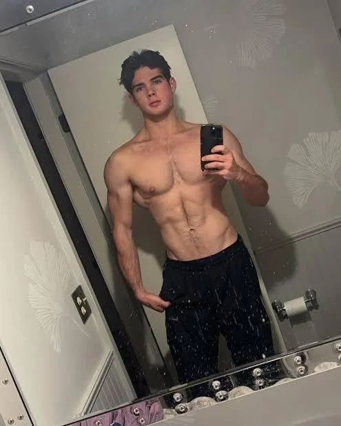 A young man with dark hair taking a mirror selfie in a bathroom. He is shirtless, wearing black shorts, and has a muscular build with defined abs and arms.