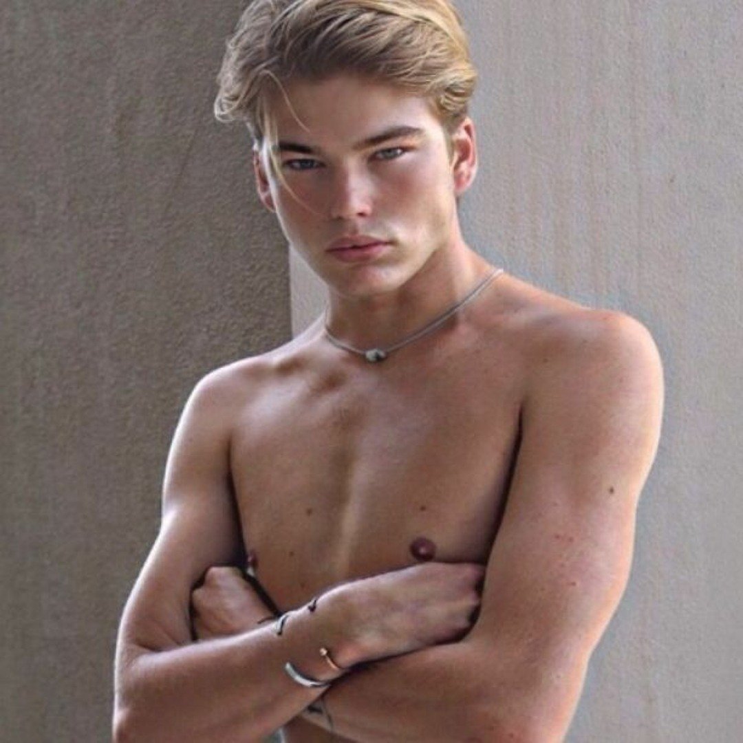 A shirtless young man with blond hair, crossed arms, and a serious expression, standing against a plain background.