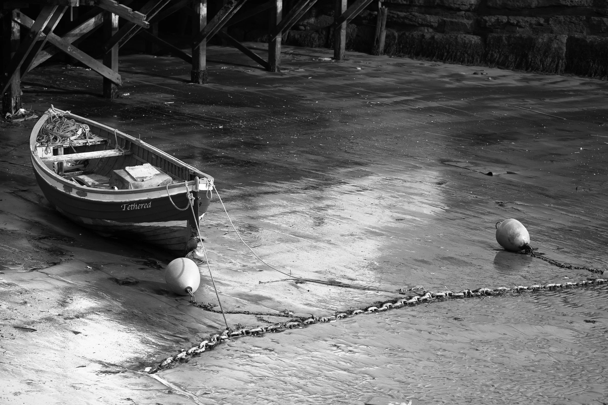 Black-and-white photograph of a small boat secured by ropes and chains in shallow water near a wooden structure at low tide.