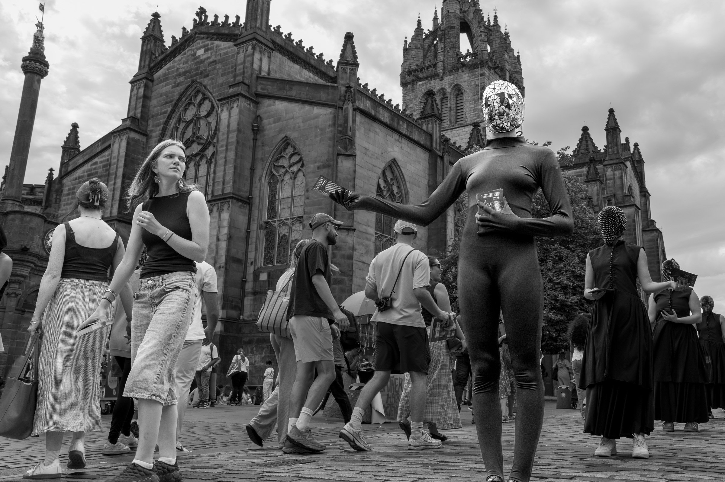 Parliament Square, Edinburgh, during Festival, with St Giles' Cathedral behind. A street performer in black bodysuit stands, head covered by a mask of mirrored fragments, holds out a flyer. Another person looking at first passes by.
