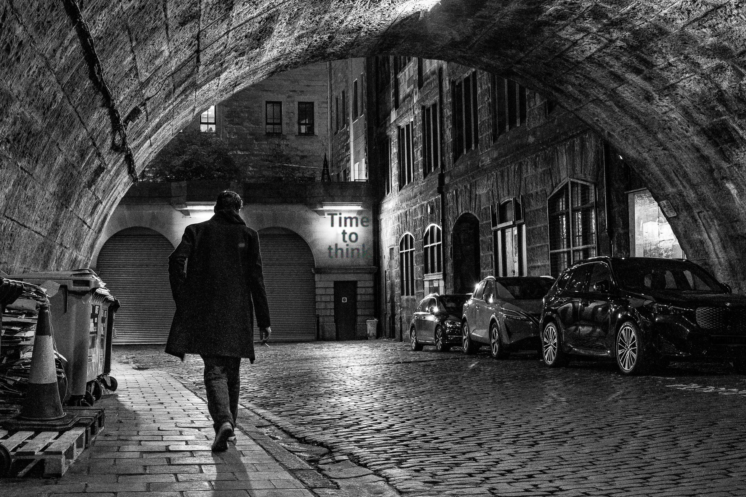 Black-and-white photograph of a person walking alone through a stone archway at night, with parked cars lining a narrow cobbled street.