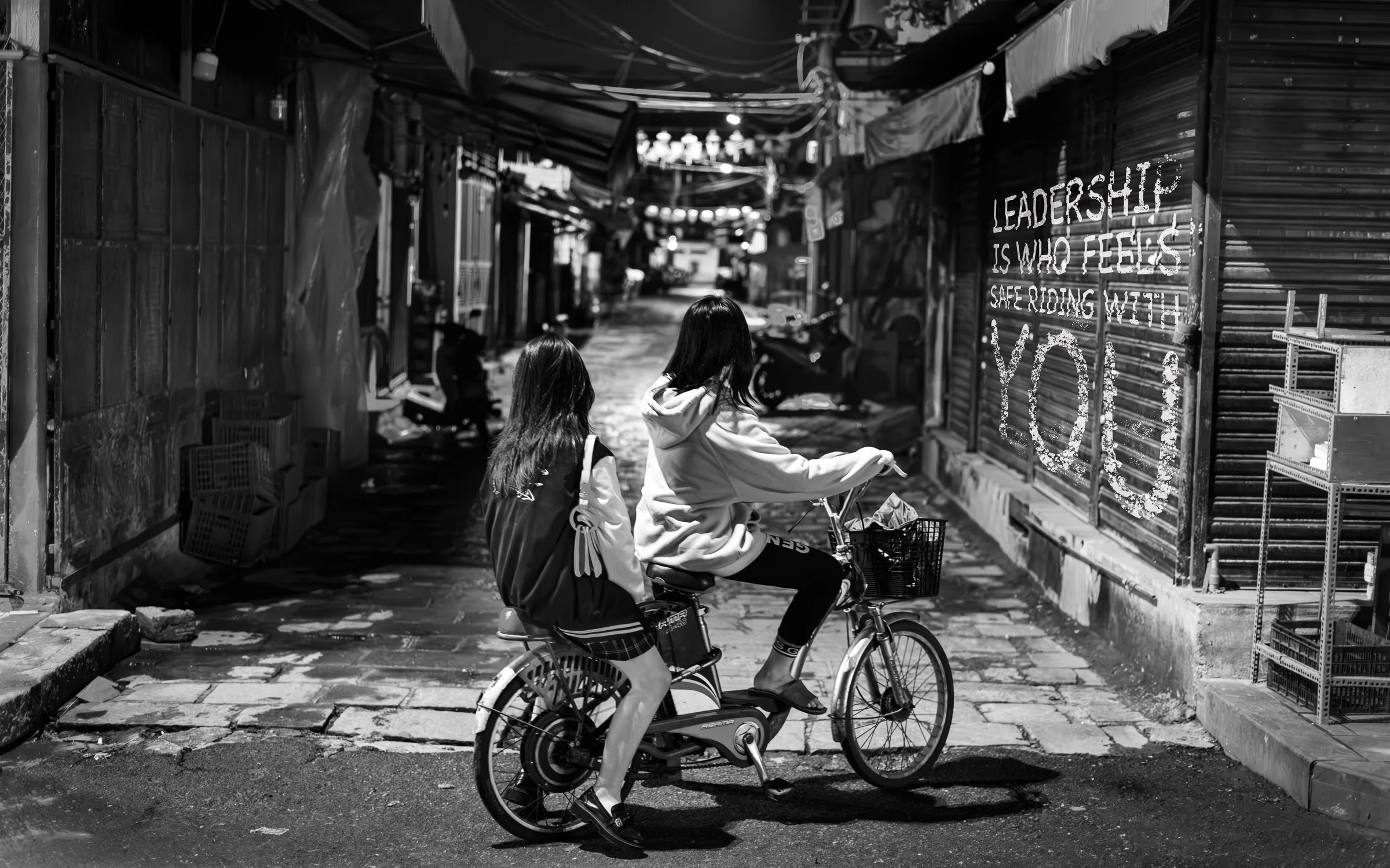Two young girls riding a bicycle together move through a narrow, graffiti-lined alleyway on a city street in black and white.