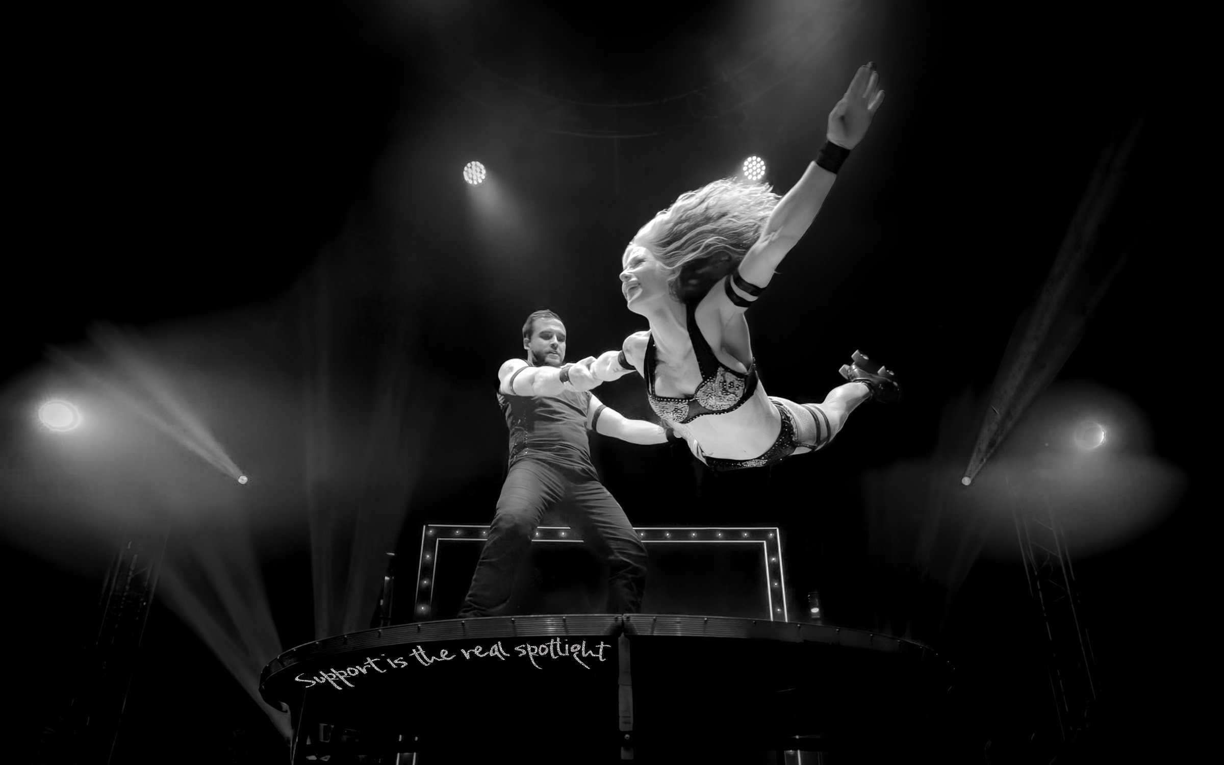 A performer is lifted into the air during a live show, with a man supporting her from below on a platform that has the phrase 'Support is the real spotlight' written on it, in an artistic black-and-white photo.