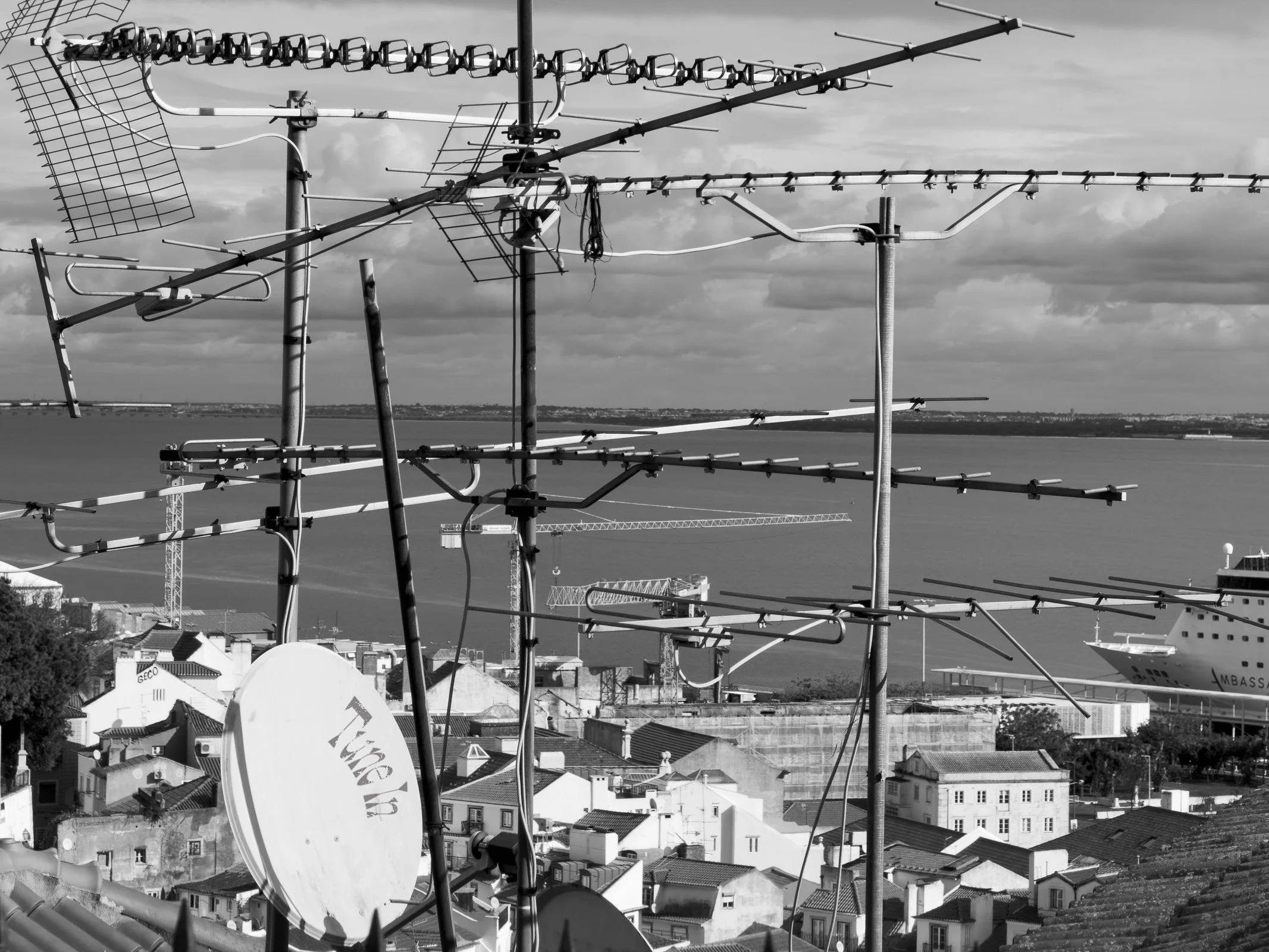 A tangle of television aerials and a satellite dish dominate the rooftops of Lisbon, with the Tagus river, construction cranes, and a cruise ship visible in the background beyond the clutter of signal receivers.
