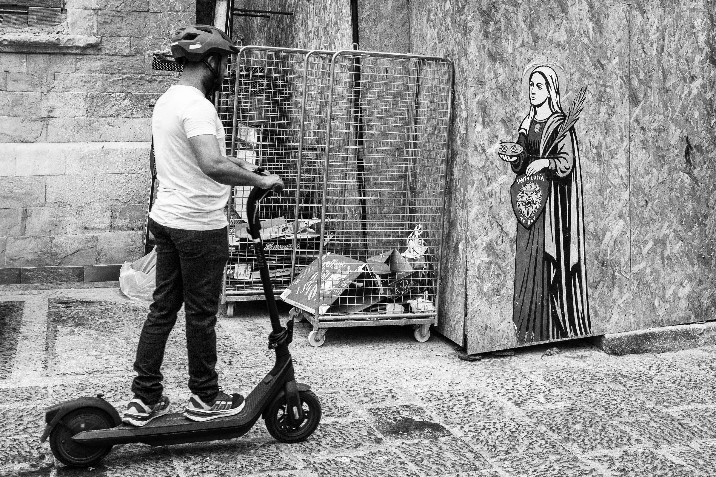 A man on an electric scooter passes a vibrant street mural of Santa Lucia painted on hoarding next to a cage of urban debris, the ancient image of the patron saint of sight unseen as he rides by.