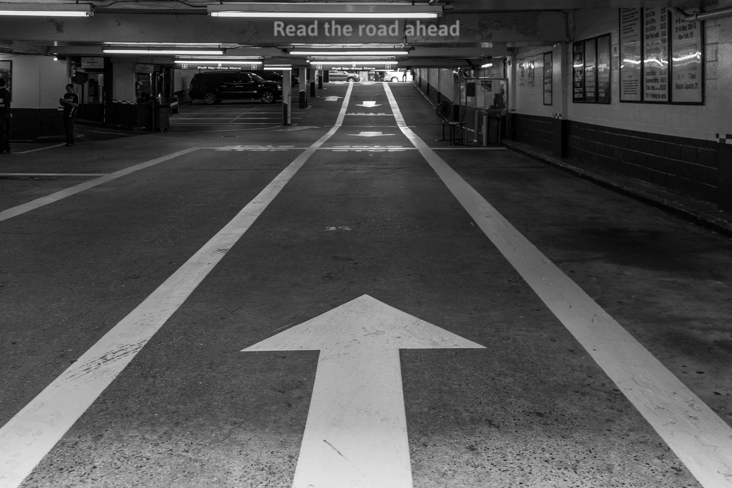 Entrance to an underground parking garage with white directional arrows on the ground pointing forward. A sign overhead reads "Read the road ahead." On closer scrutiny arrows approach driver giving concern for collision.