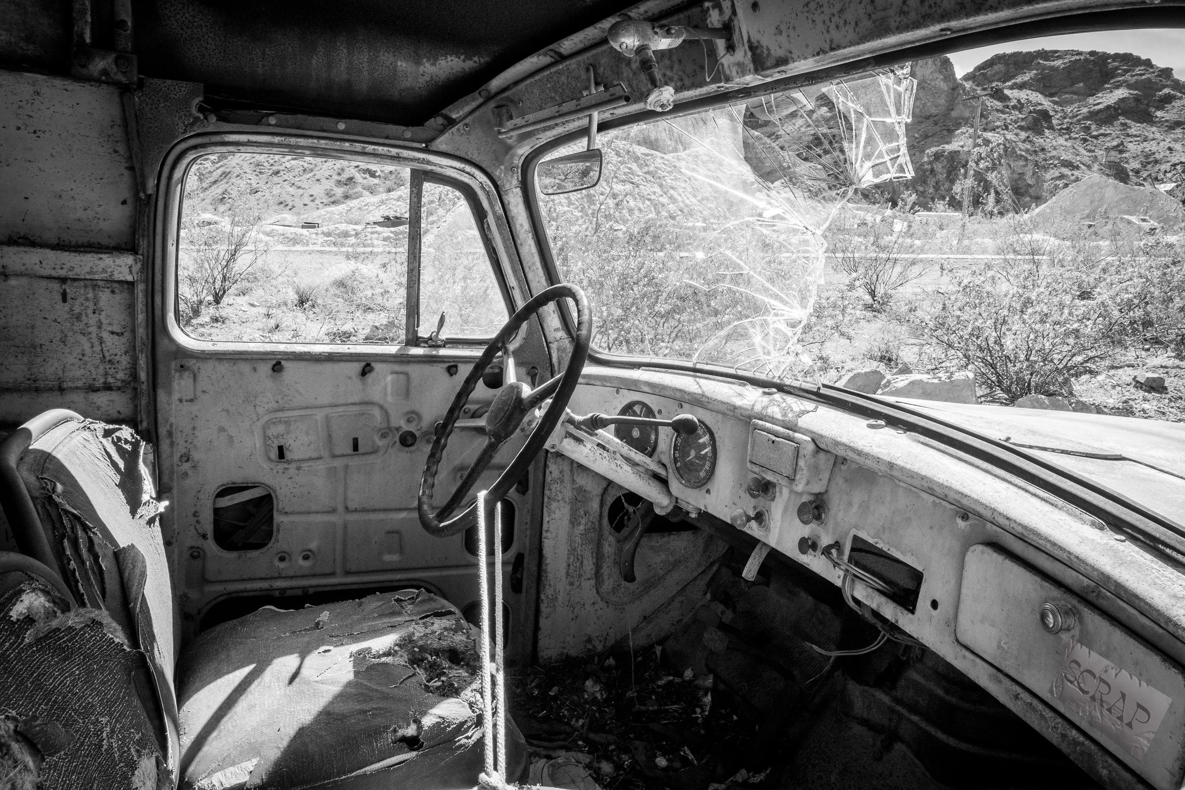 The interior of an abandoned vintage truck, its windscreen shattered into a spider-web pattern, the torn seat and rusted dashboard framing a view of desert mountains and scrubland beyond.