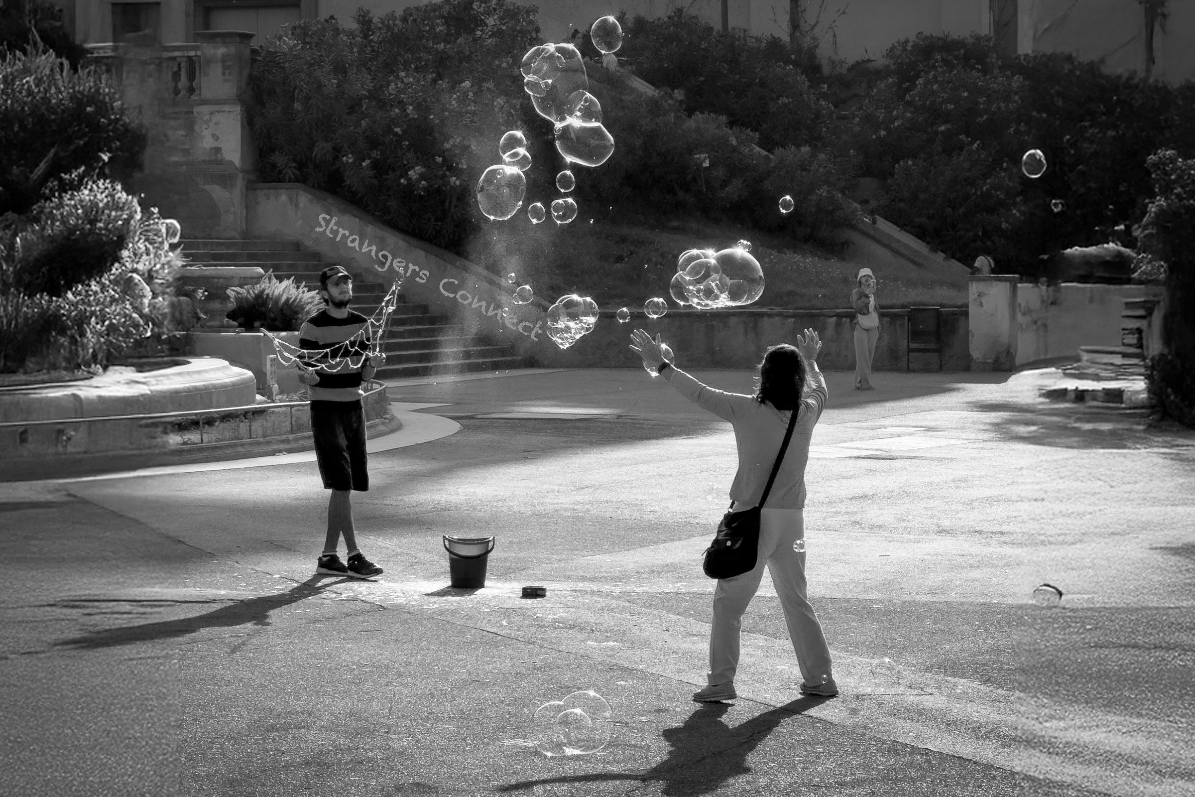 Black-and-white photograph of two people facing each other in an open public space, creating large soap bubbles between them while others stand nearby.