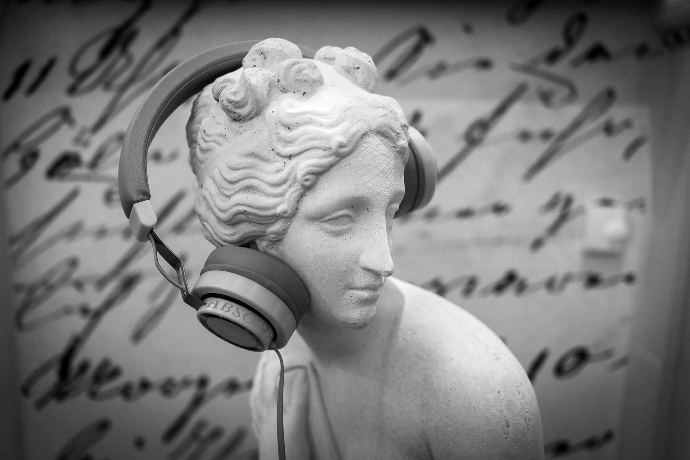 A classical marble bust wearing modern headphones, with handwritten script visible in the background, the juxtaposition of ancient contemplation and contemporary consumption.