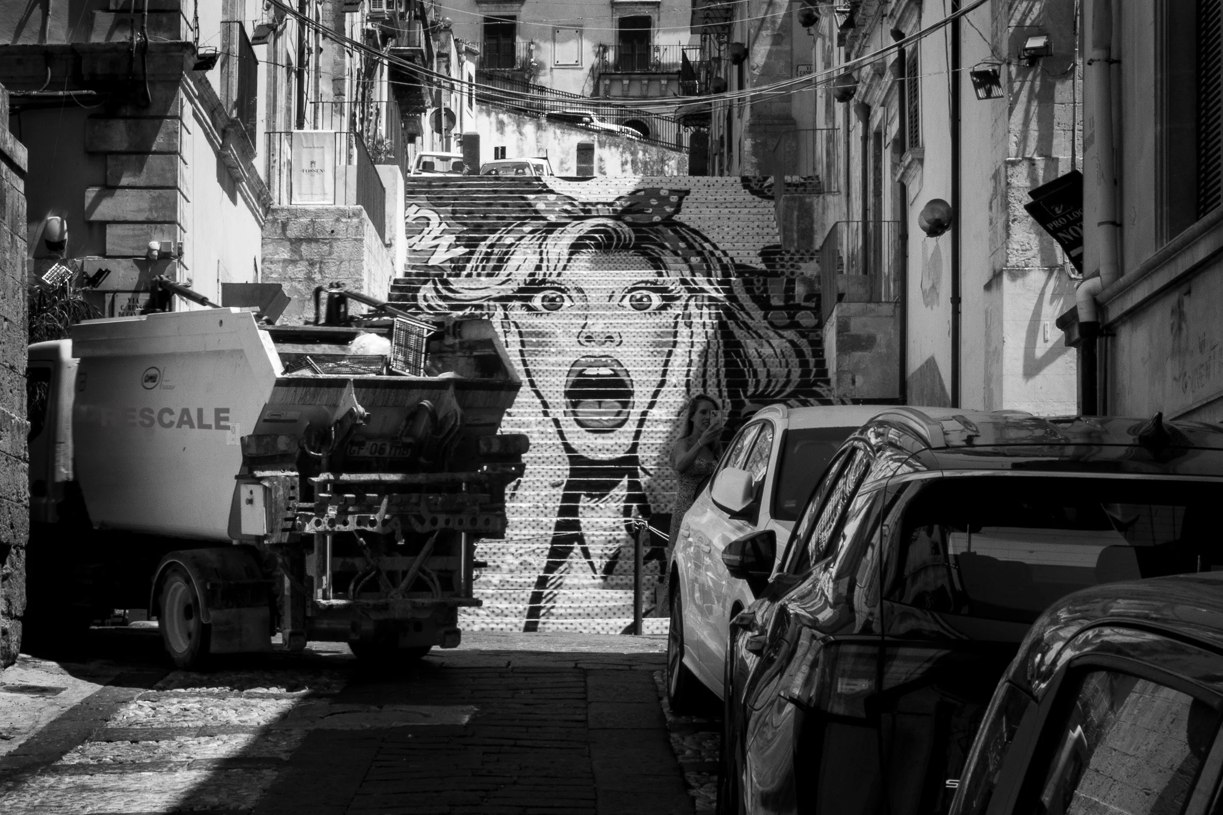 A large pop art mural riffing on Edvard Munch’s The Scream painted across a staircase in a narrow Sicilian street, dwarfed by parked cars and a passing truck, the figure’s expression of existential anguish ignored by the ordinary activity around it.