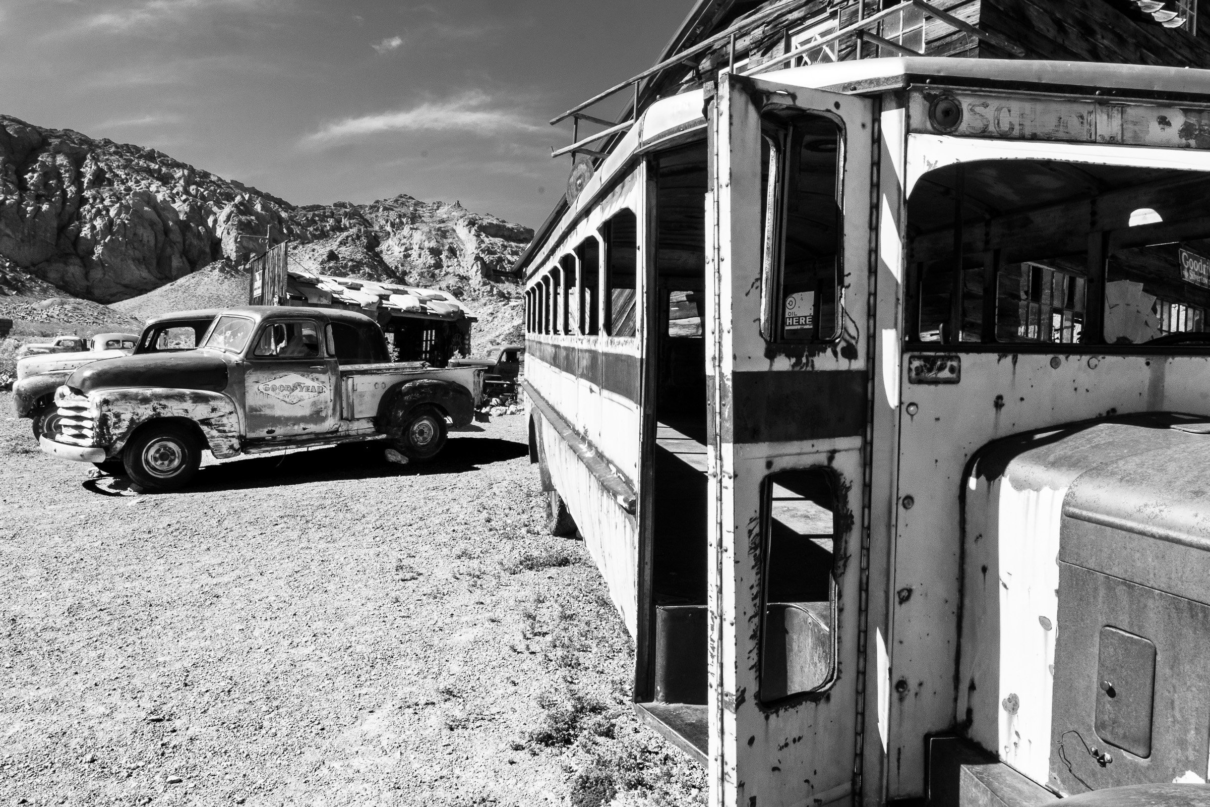 Abandoned vintage vehicles, including a rusted bus with its door left open and a Goodyear pickup truck, sit decaying in a desert ghost town beneath rugged mountains.