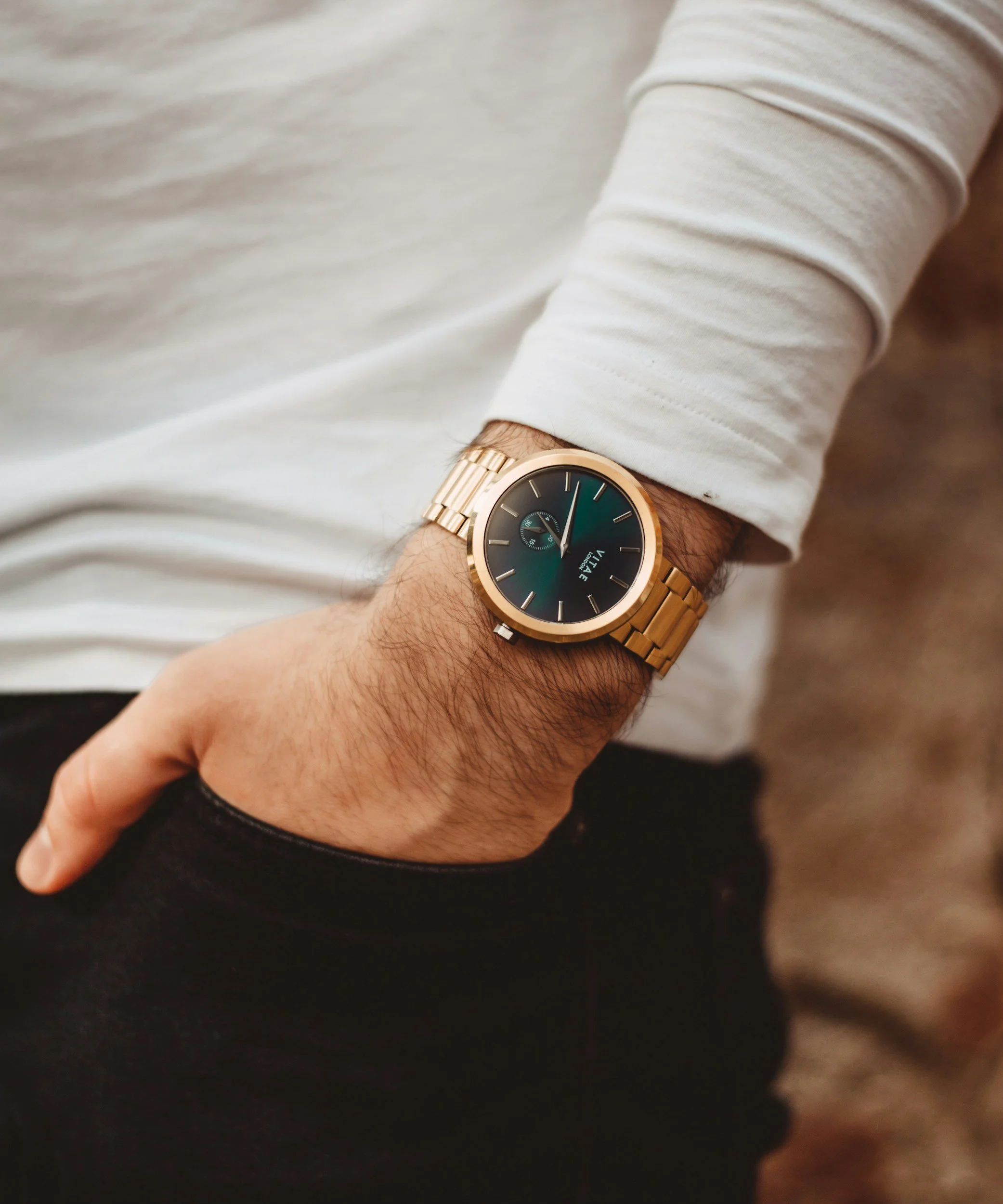 A man wearing a white long-sleeve shirt and a gold wristwatch with a black and green watch face, resting his hand in his pocket.
