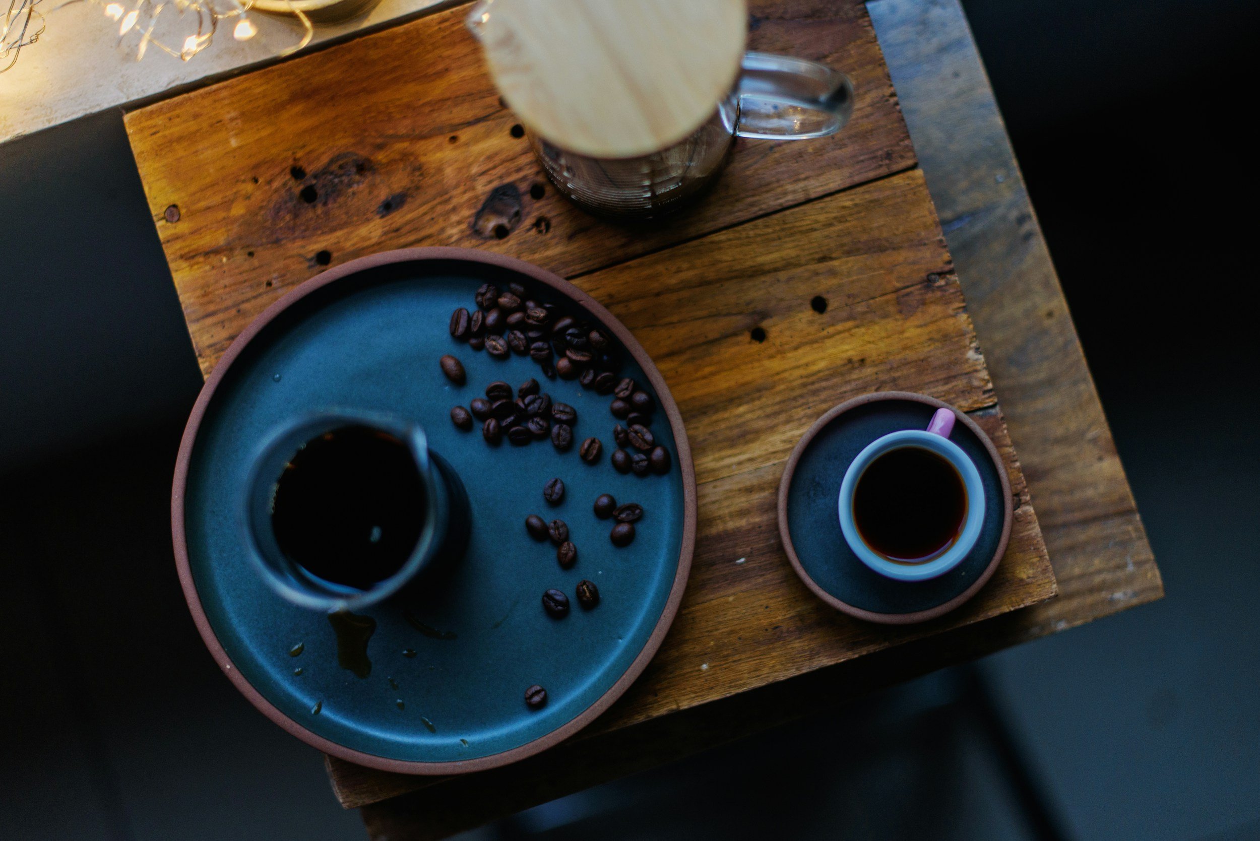Top-down view of a rustic wooden table with a blue plate holding scattered coffee beans, a mug of black coffee, and a small cup of black coffee on a matching saucer.