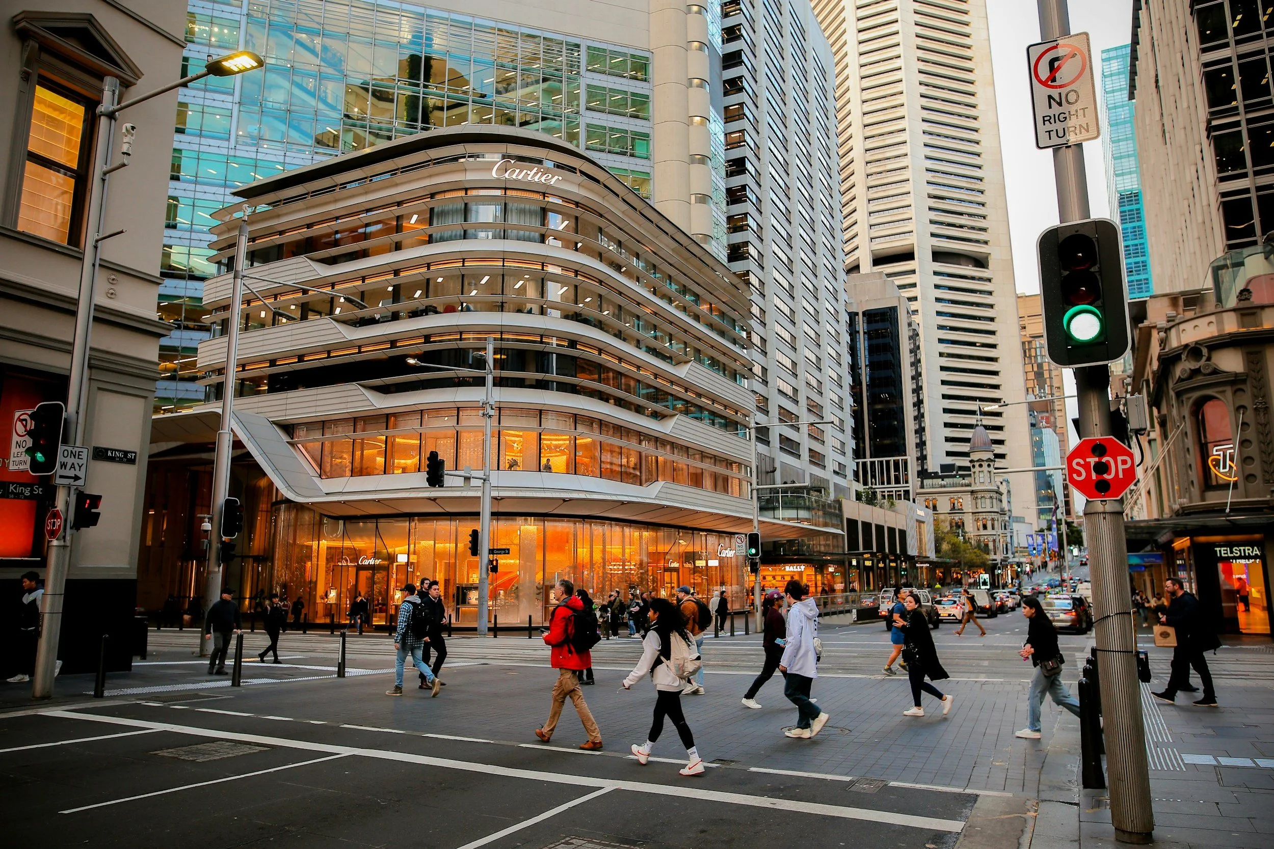 Street scene in an urban area with tall buildings, a luxury clothing store, and a busy crosswalk with pedestrians. Traffic lights and street signs are visible.