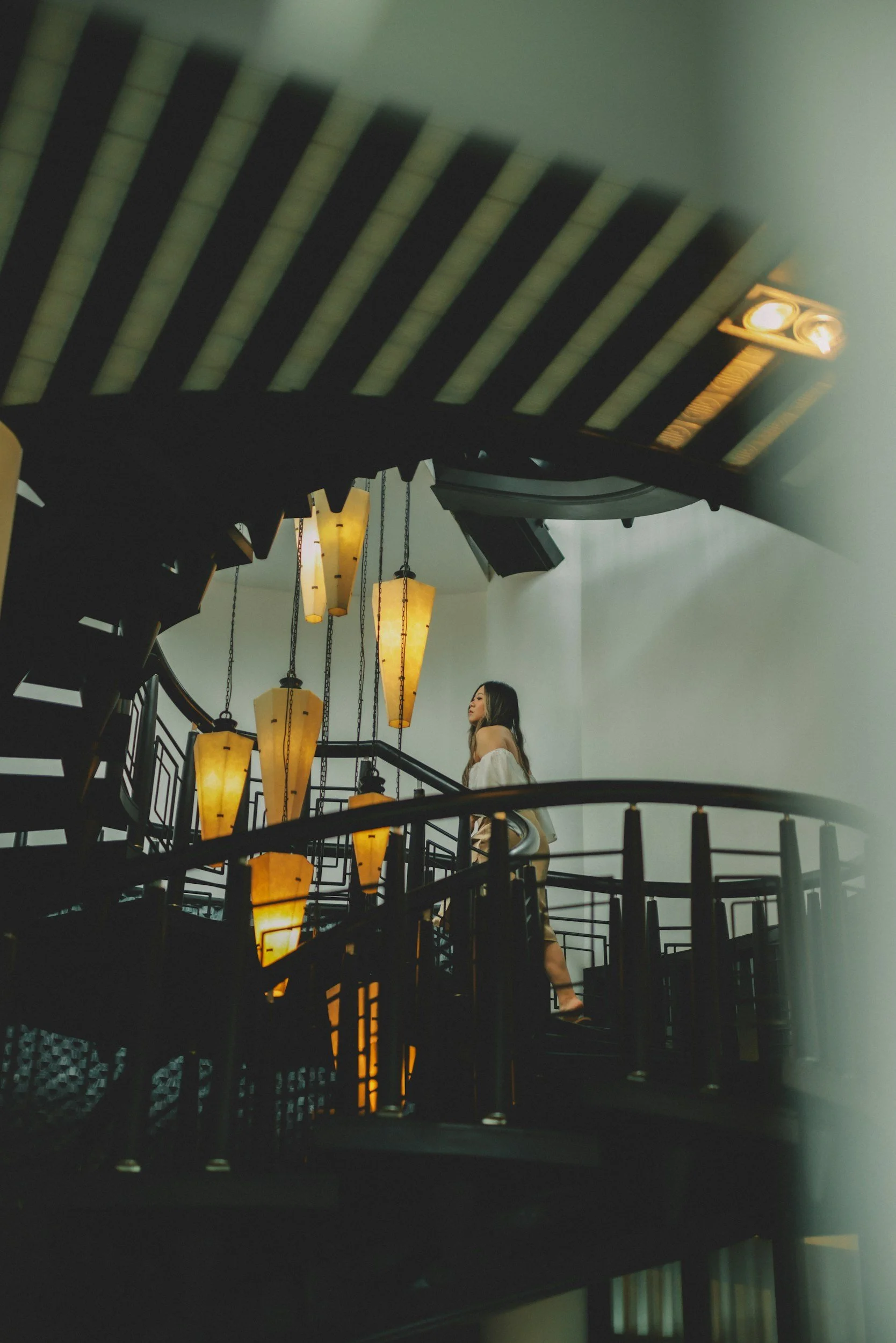 A woman standing on a spiral staircase near hanging lanterns in an indoor setting.
