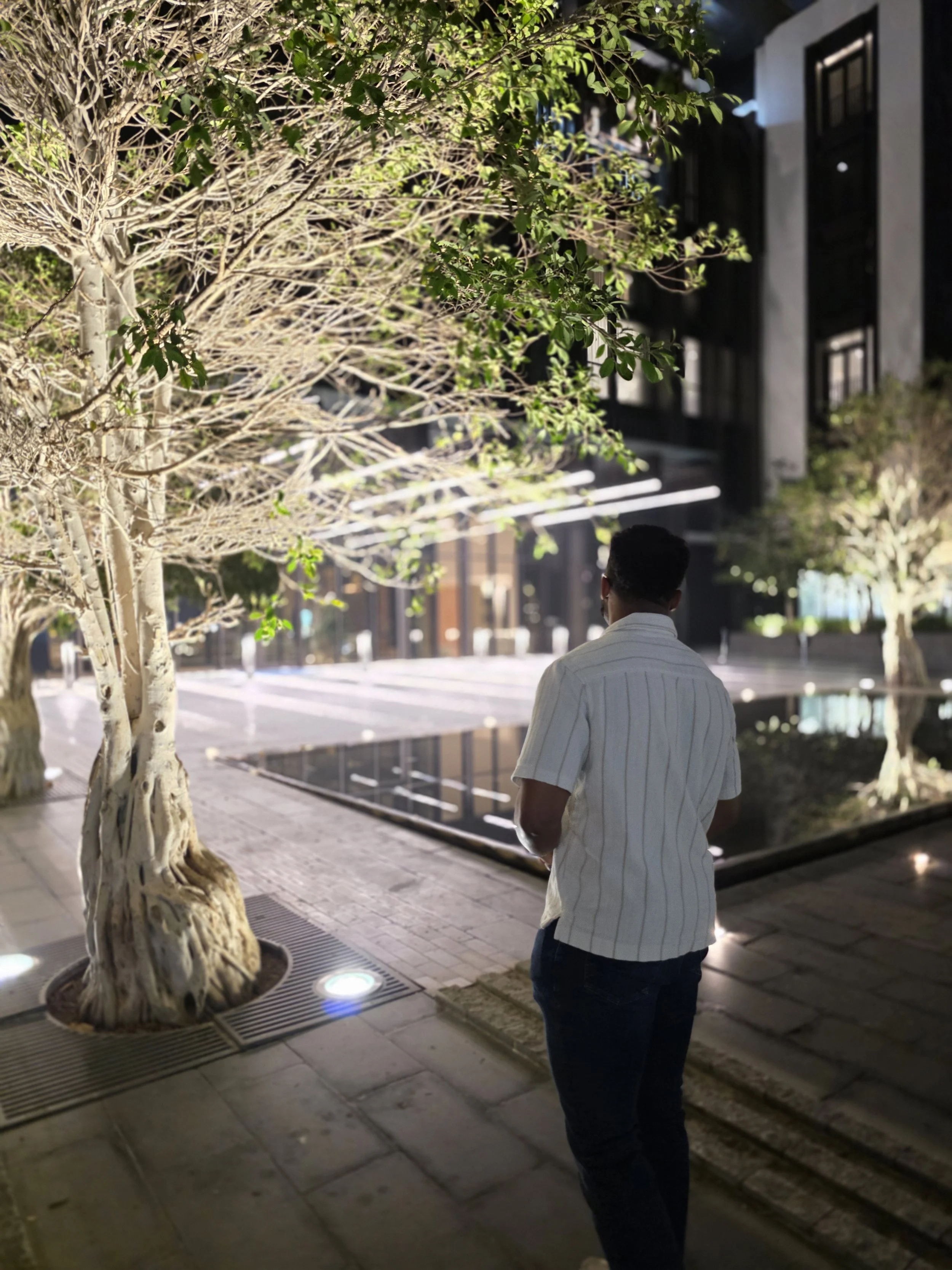 A man with short hair, wearing a white shirt and dark jeans, stands on a lit sidewalk at night near a tree with green leaves and a water feature in an urban environment.