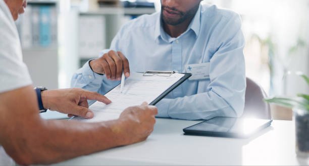 A person in a white shirt points to a document on a clipboard held by another person in a blue shirt, in a well-lit office setting.