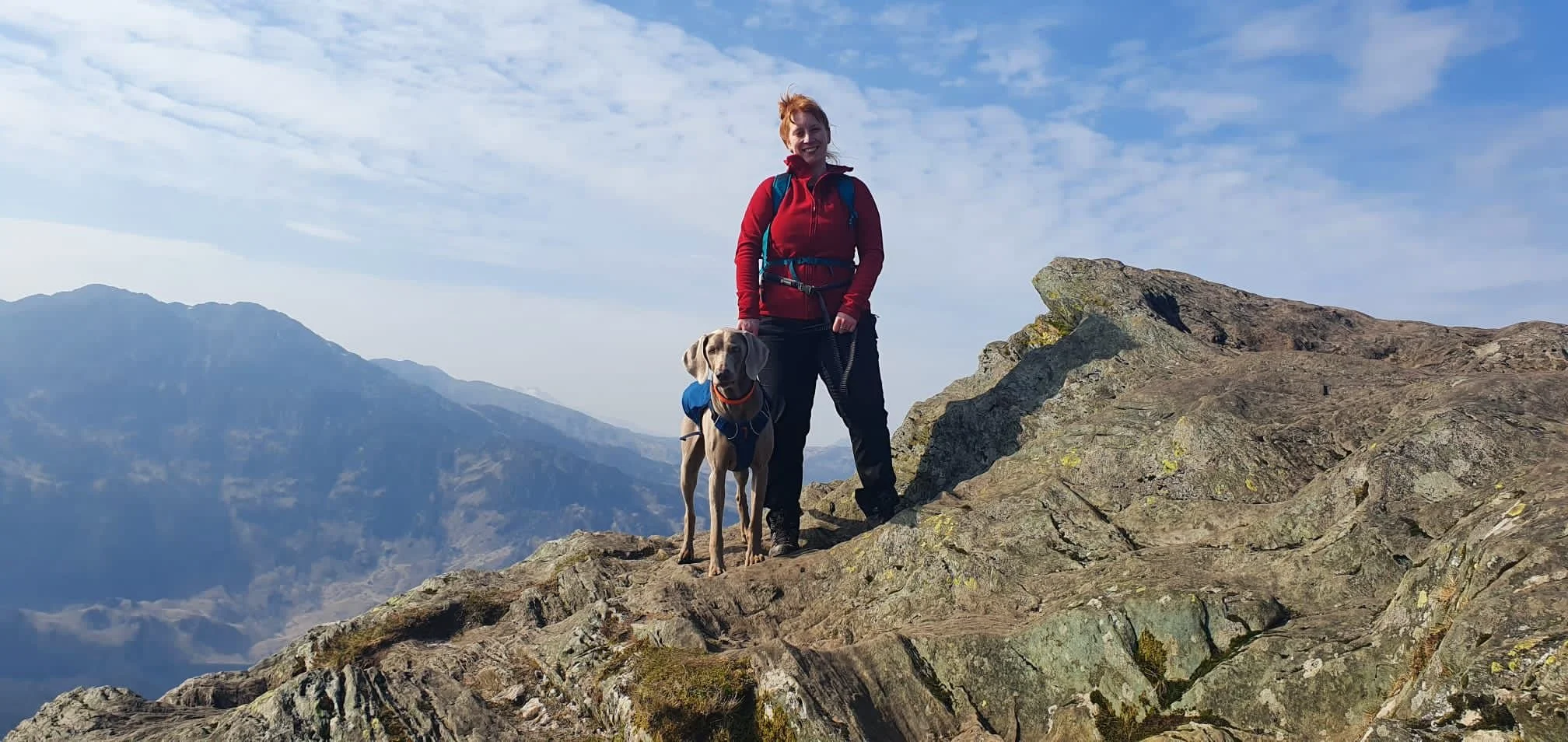A woman in red jacket hiking with her dog on a rocky mountain summit, with mountain ranges and a blue sky with clouds in the background.
