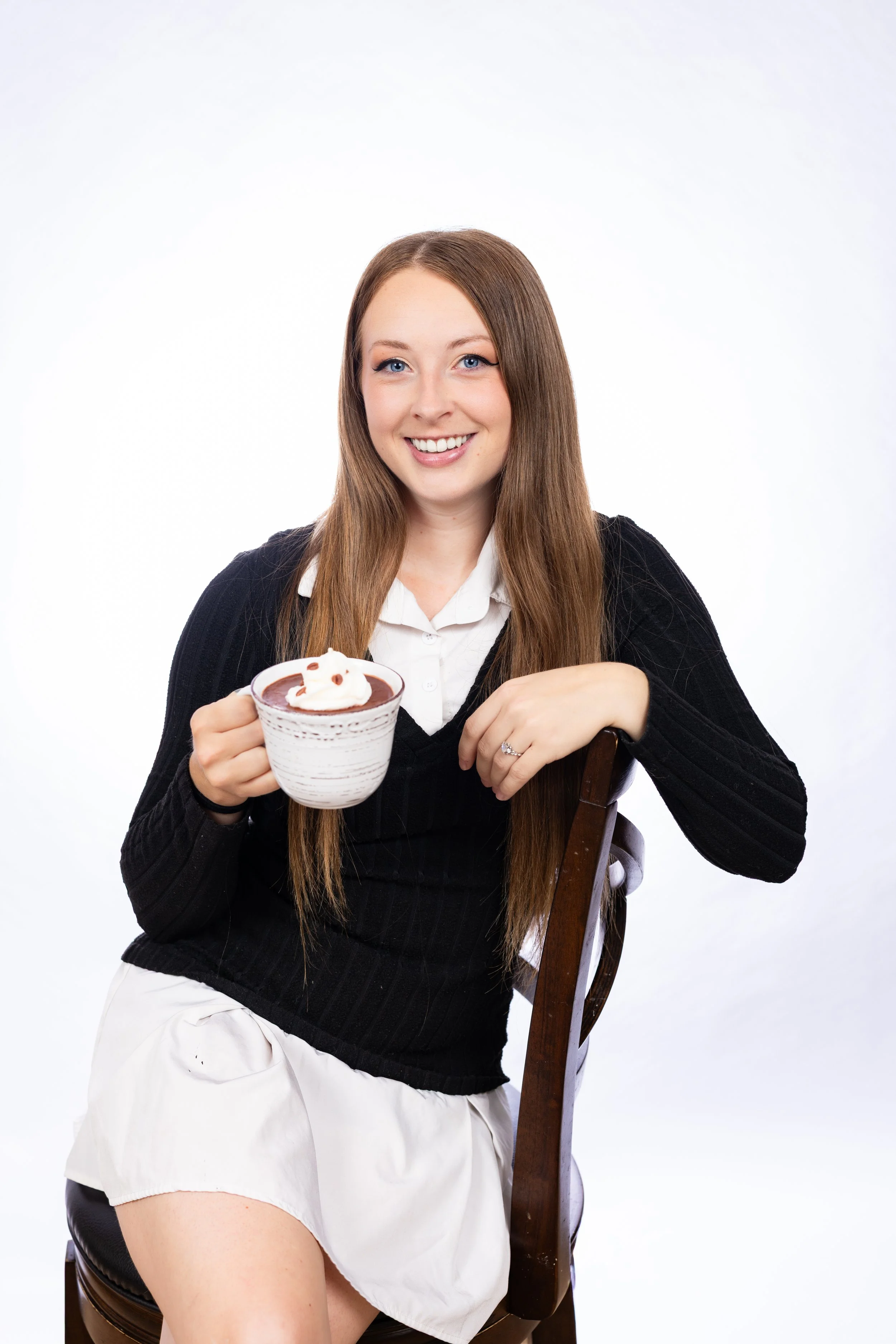 A woman with long brown hair, wearing a white blouse and black cardigan, smiling and holding a bowl of chocolate pudding with whipped cream, sitting on a wooden chair against a plain white background.