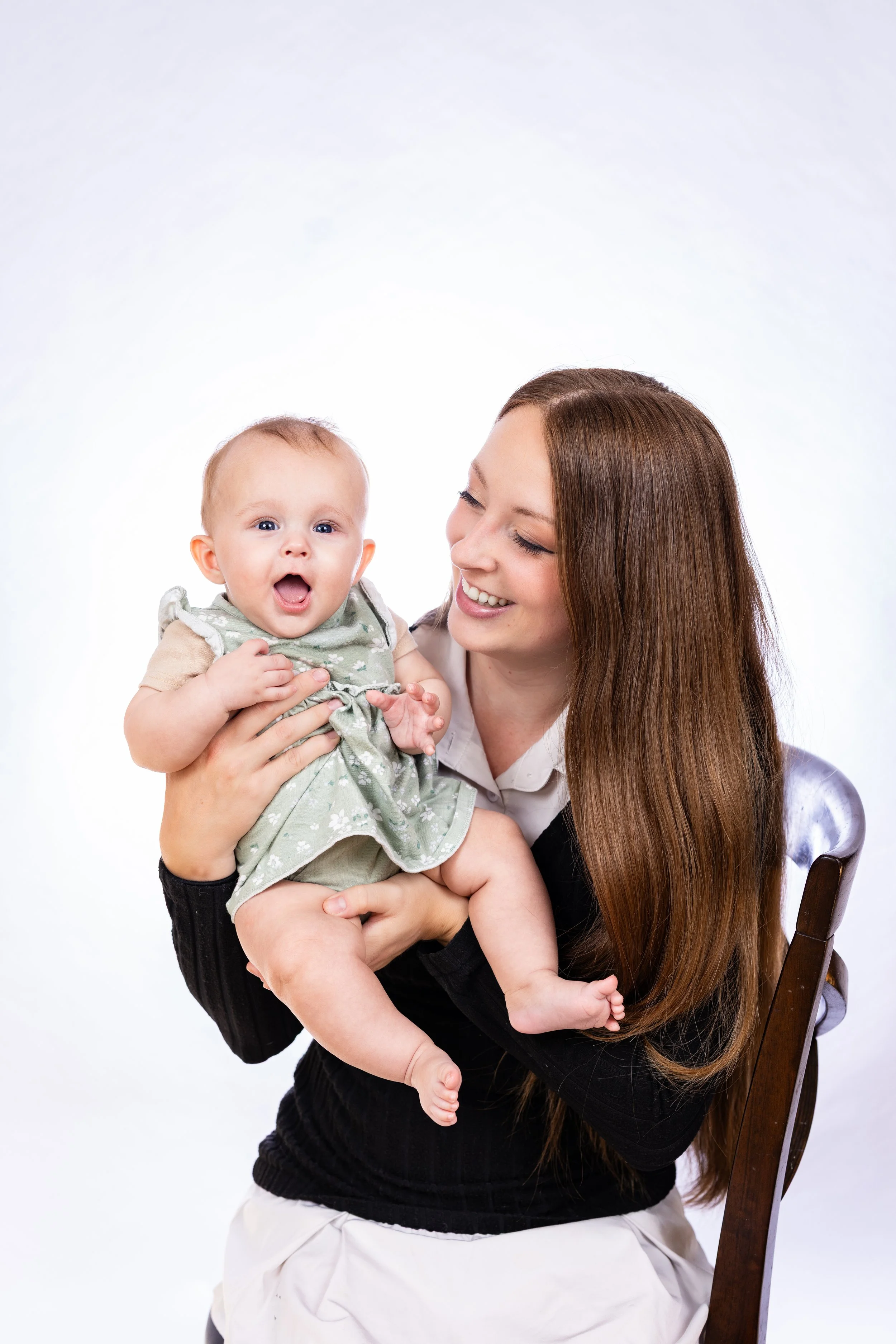 A woman with long brown hair smiling and holding a happy baby with blue eyes and blond hair, against a white background.