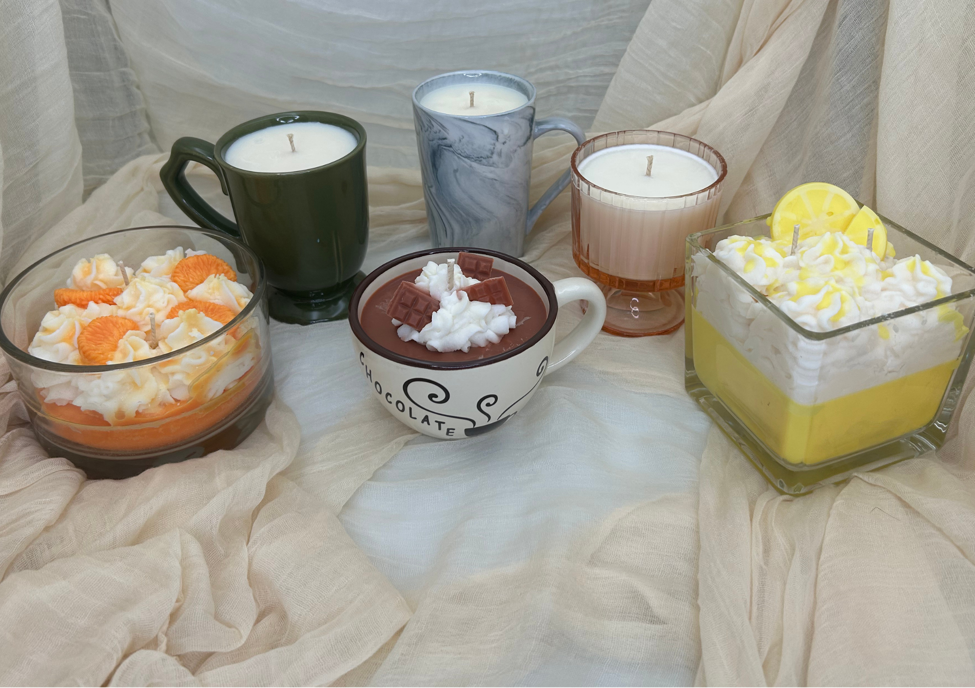 A collection of candles in different holders and colors, including a marble-patterned mug and a glass container with yellow and white candles, displayed on a cream fabric background.