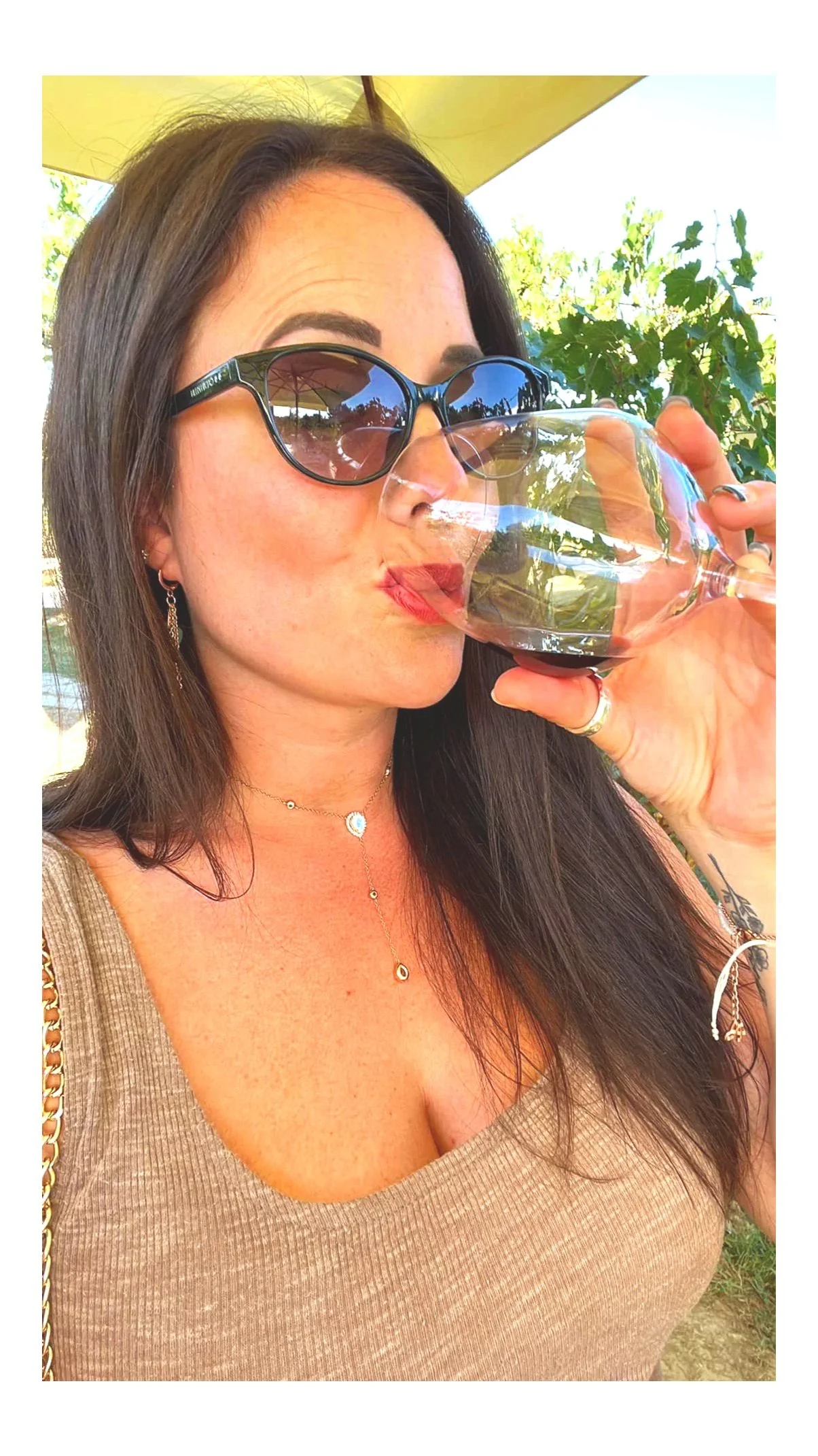 Woman with long dark hair wearing sunglasses, jewelry, and a beige top drinking red wine outdoors on a sunny day.