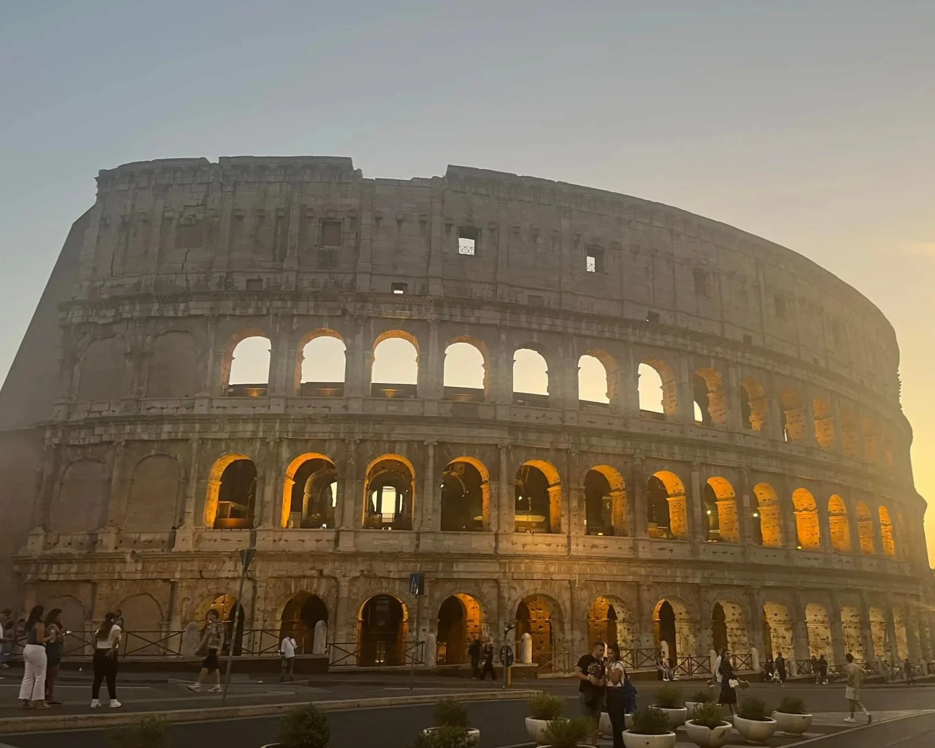 The Colosseum in Rome illuminated at sunset with people walking in front.