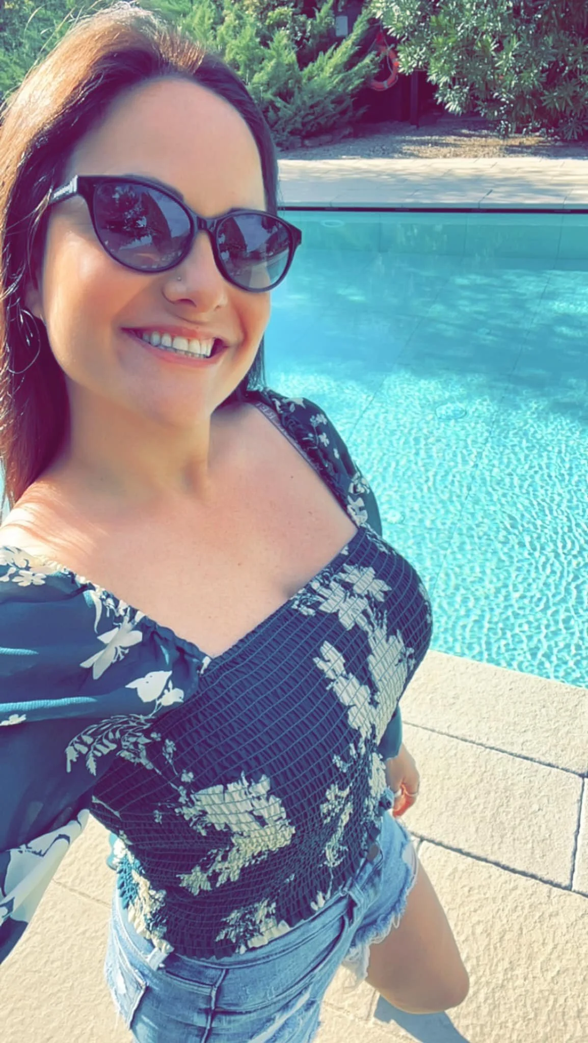 Woman smiling by a swimming pool, wearing sunglasses, a floral top, and denim shorts, with greenery in the background.