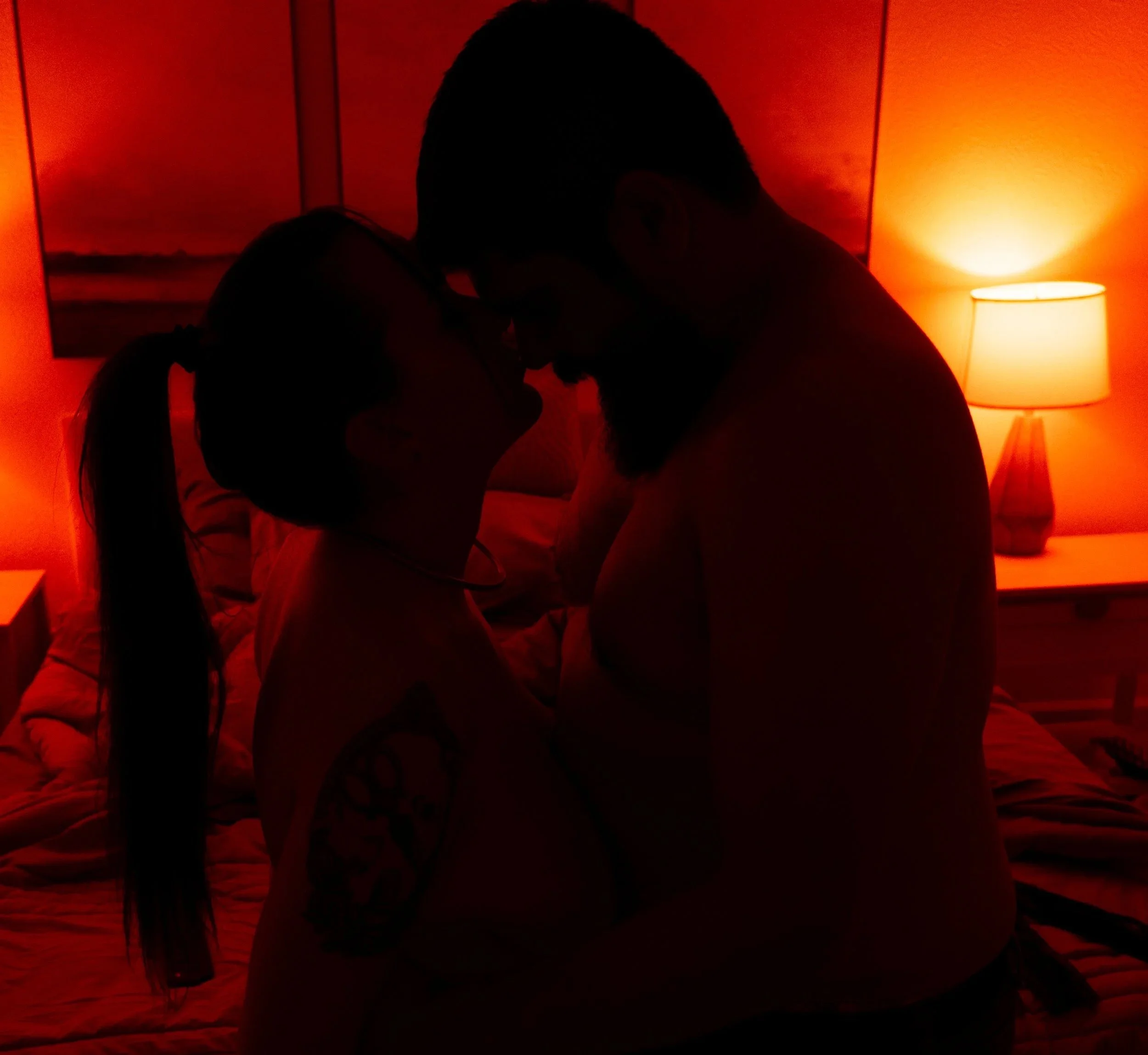 Silhouetted couple in an embrace in a dimly lit bedroom with warm lighting from a bedside lamp.
