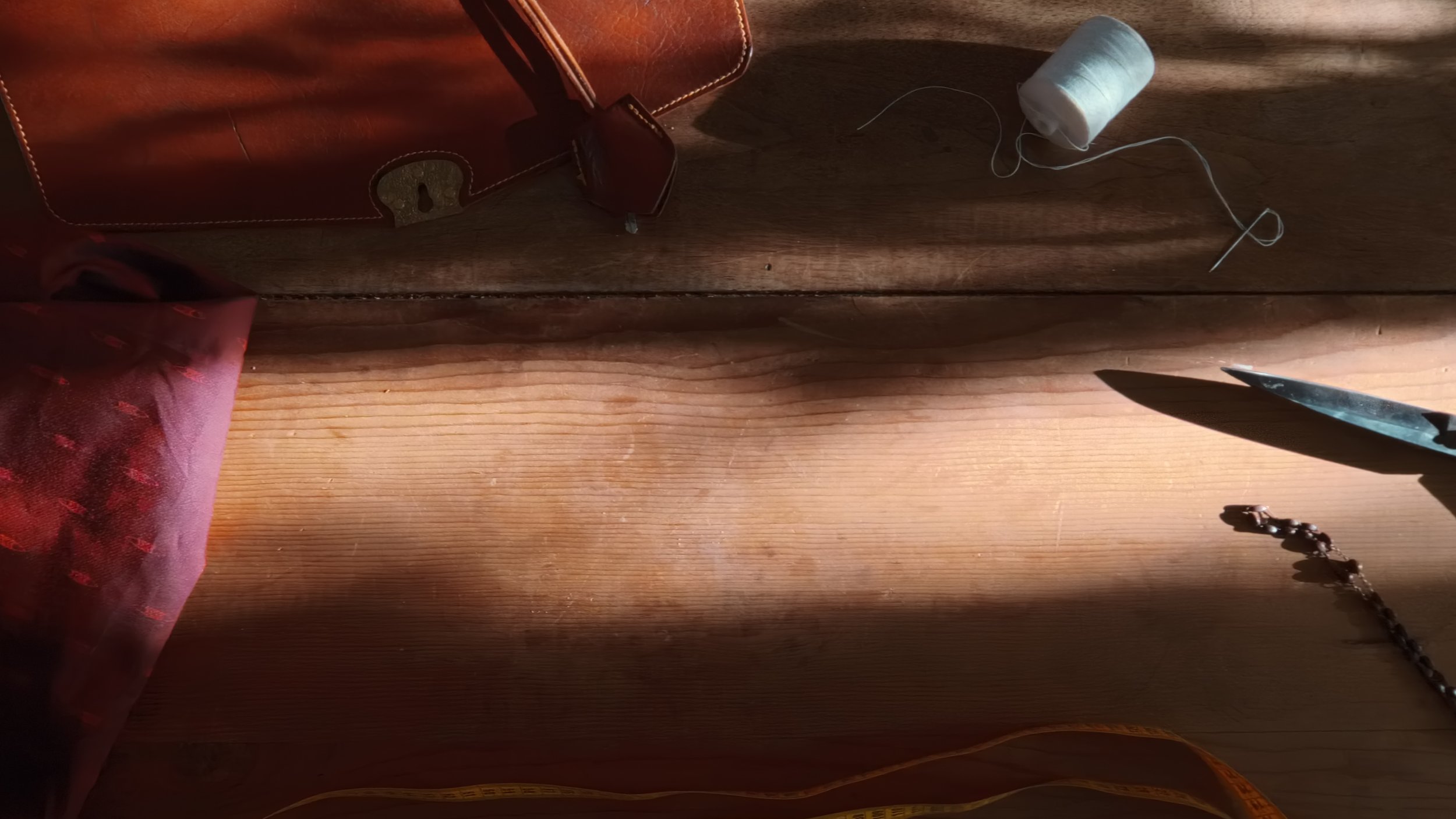 Une boîte en cuir marron ouverte sur une table en bois, un fil de couture blanc, une bobine de fil blanc, un ciseau, et un bracelet en perles