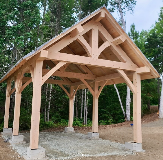 A wooden pavilion under construction in a forested area with trees in the background, supported by concrete footings.
