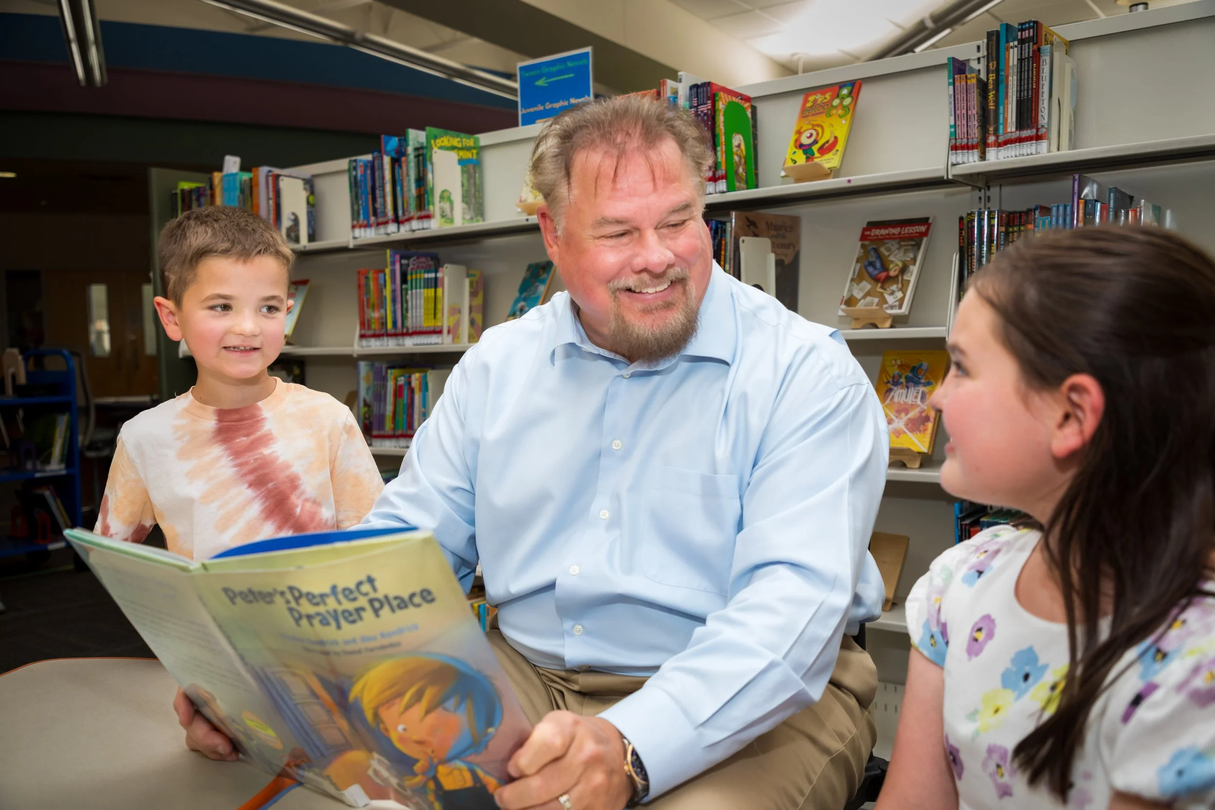 John Bachman sitting in a library reading a book titled 'Perfect Prayer Place' to two smiling children, one girl with dark hair and a floral shirt, and one boy with short hair and a tie-dye shirt.