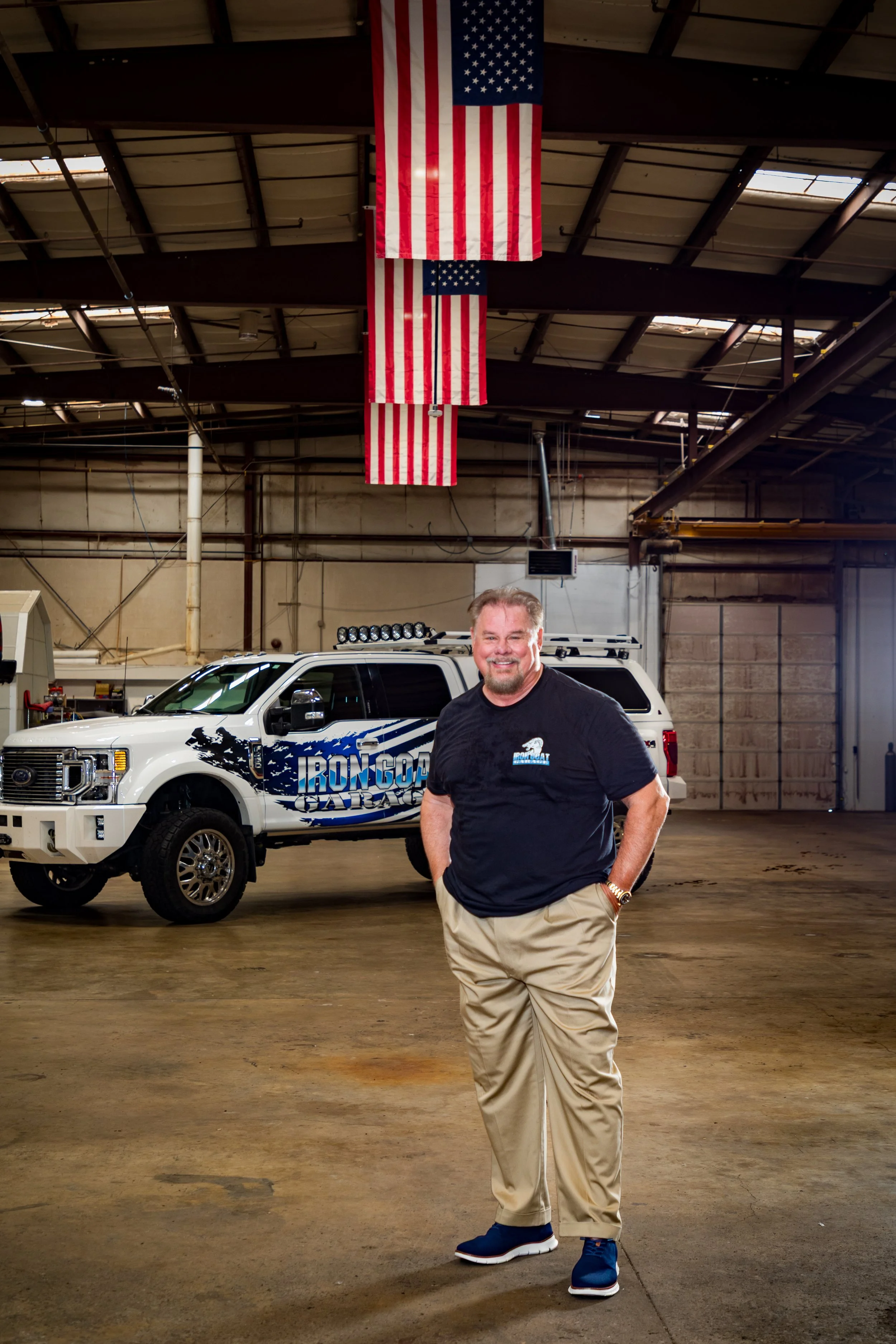 John Bachman smiling, posing with hands in pockets, standing inside Iron Goat Garage next to a white utility vehicle, under hanging American flags.
