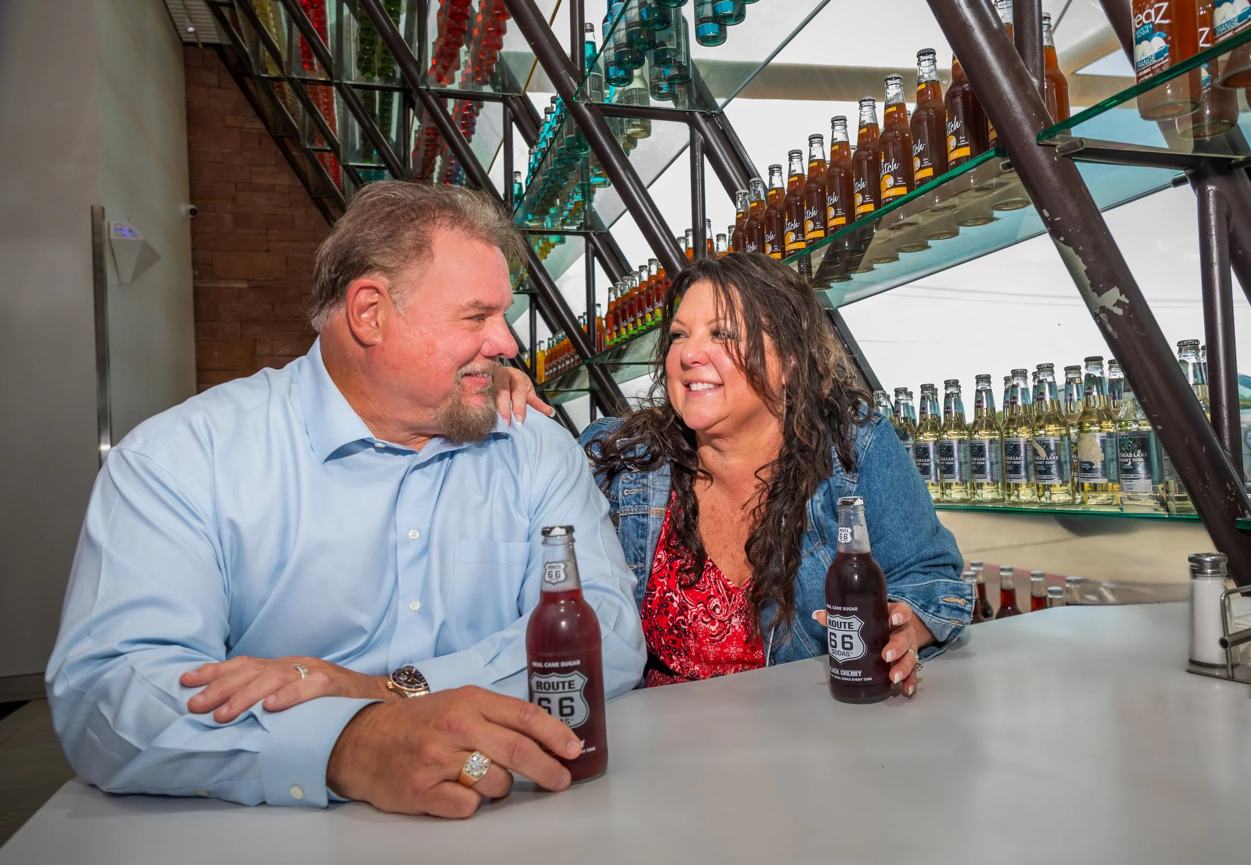 John and Sheri Bachman sitting at a table inside Pop's, smiling and looking at each other, each holding a Route 66 soda bottle. The bar has glass shelves displaying various colorful bottles and cans.