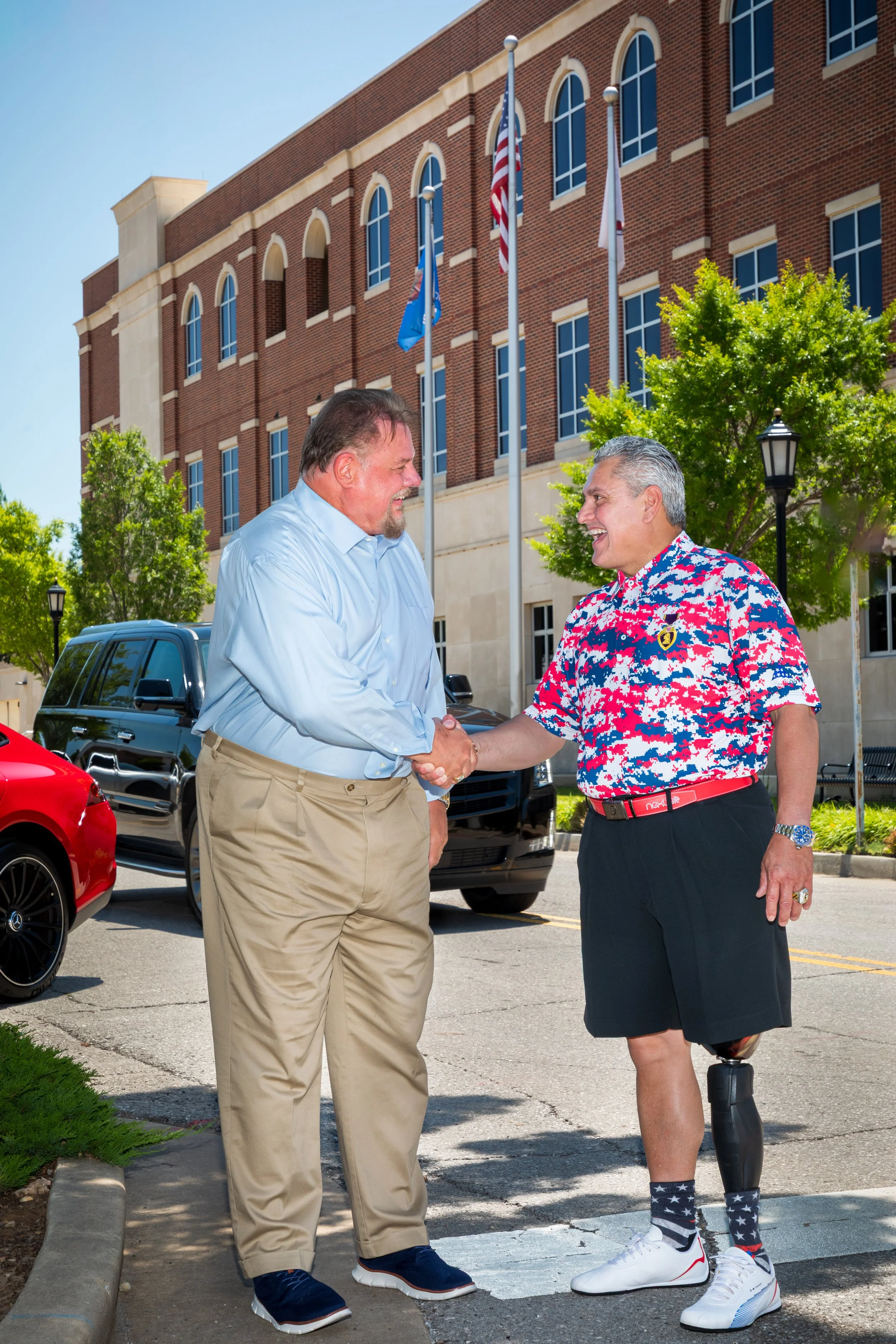 John Bachman shaking hands outside a building with flags, one in business casual and the other in a colorful shirt, shorts, and wearing a prosthetic leg.