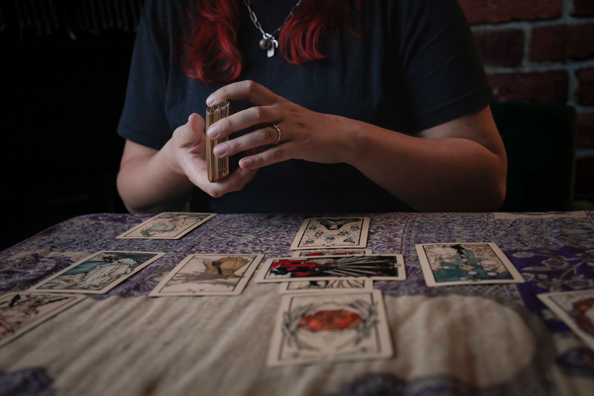 A person with red hair, wearing a black shirt, sitting at a table with a patterned cloth, holding a deck of tarot or playing cards. Several cards are laid out on the table in front of them, with a red brick wall in the background.