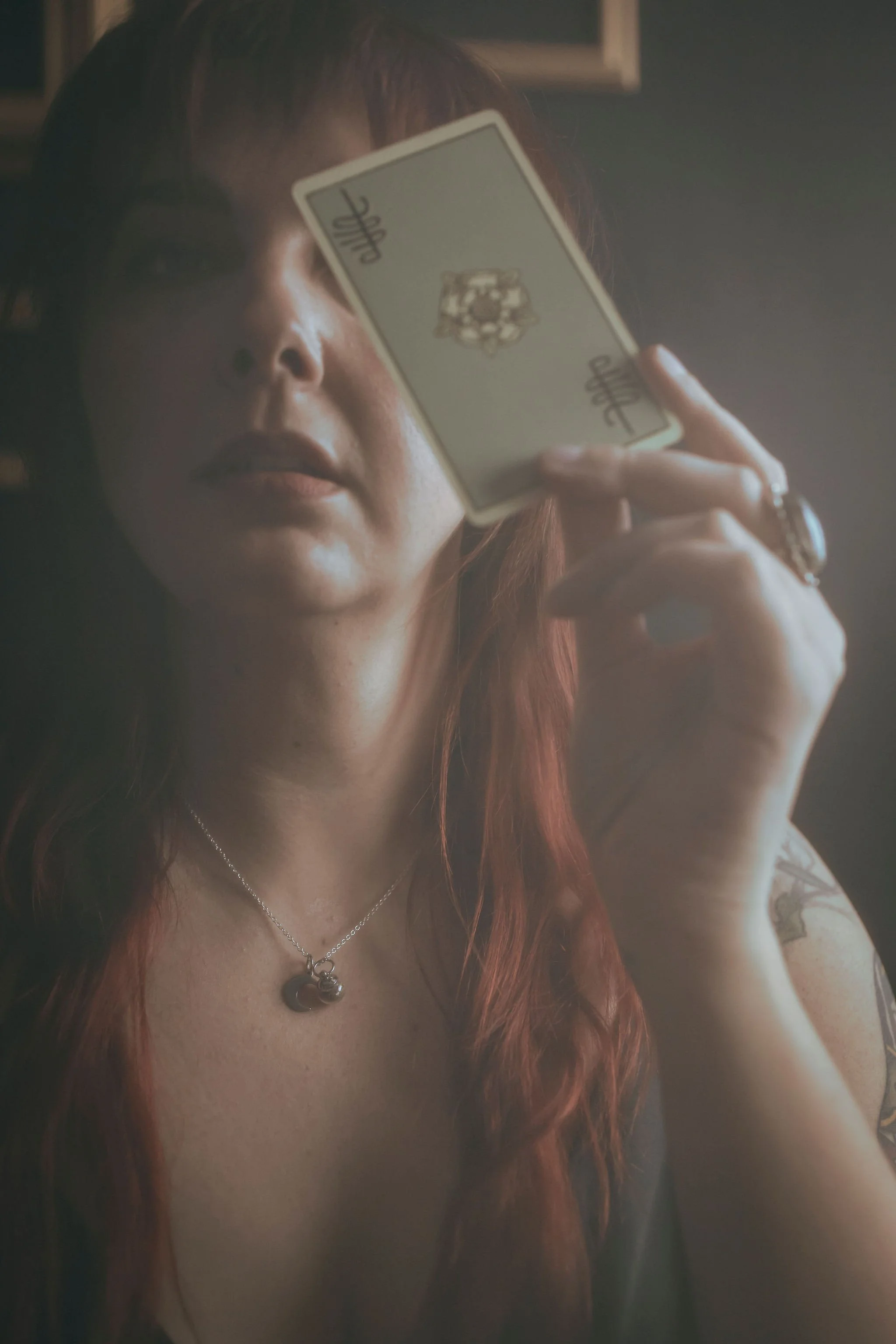 A woman with red hair holding a small mirror up to her eye, reflecting her face. She is wearing a necklace with a pendant, rings, and has a tattoo on her arm. The photo has a soft, dim lighting.