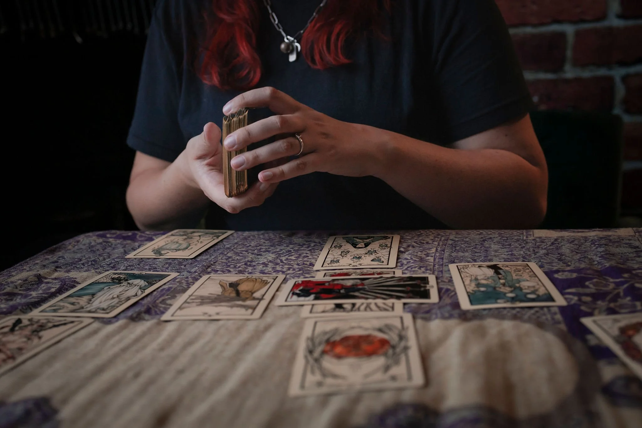 A person with red hair and jewelry sits at a table with tarot cards, holding a deck in their hands, with other tarot cards laid out on the table.