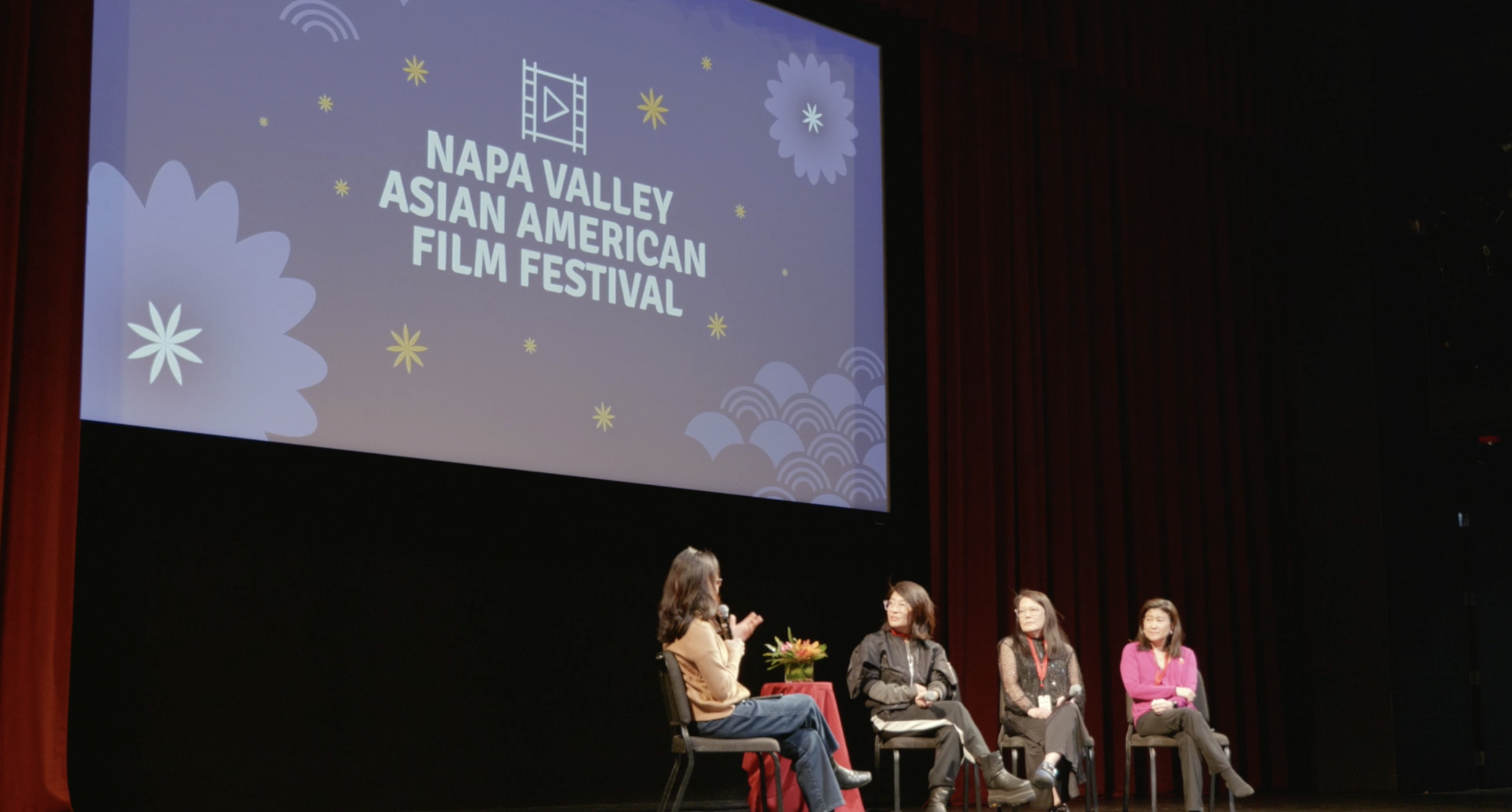 Panel discussion at Napa Valley Asian American Film Festival with four women on stage, one holding a microphone, seated in chairs, with a large screen behind displaying the festival's name and logo.