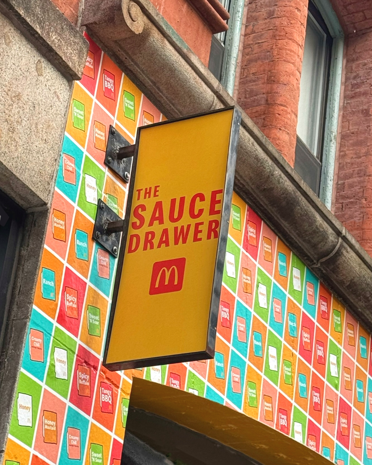 Yellow sign with red text reading "The Sauce Drawer" and McDonald's logo, mounted outside a building with a colorful menu or advertisement display in the background.