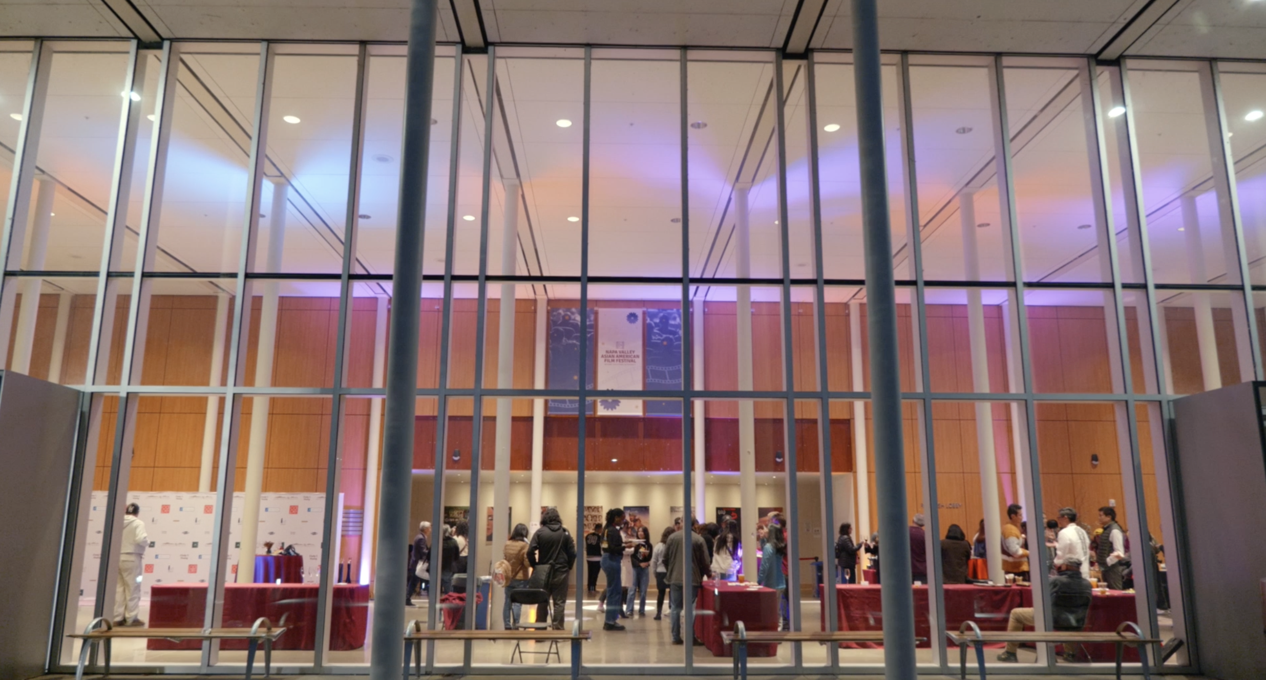 Inside a modern conference center with large glass walls, people are gathered around tables with red tablecloths, engaging in conversations and activities. The background features a banner and artwork, with warm wooden paneling and purple and blue lighting.