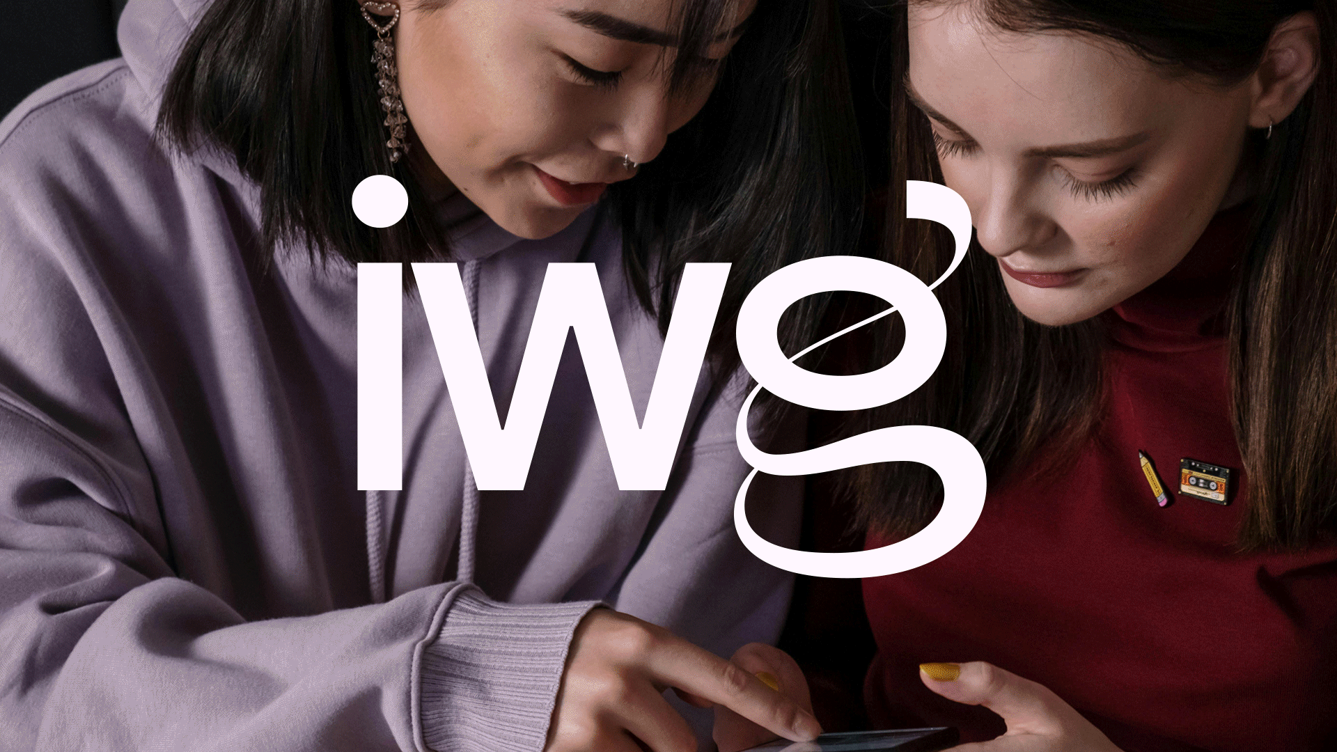 Two young women leaning close together looking at a smartphone, one wearing a lavender hoodie and jewelry, the other wearing a dark red shirt with patches, dark hair, both focused on the phone screen. IW Group Logo animation overlaid.